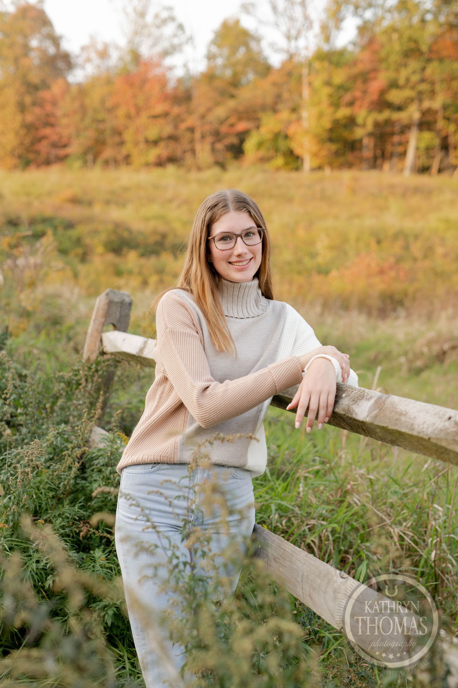 Portrait in autumn field with cozy sweater leaning against rustic wooden fence during golden hour.
