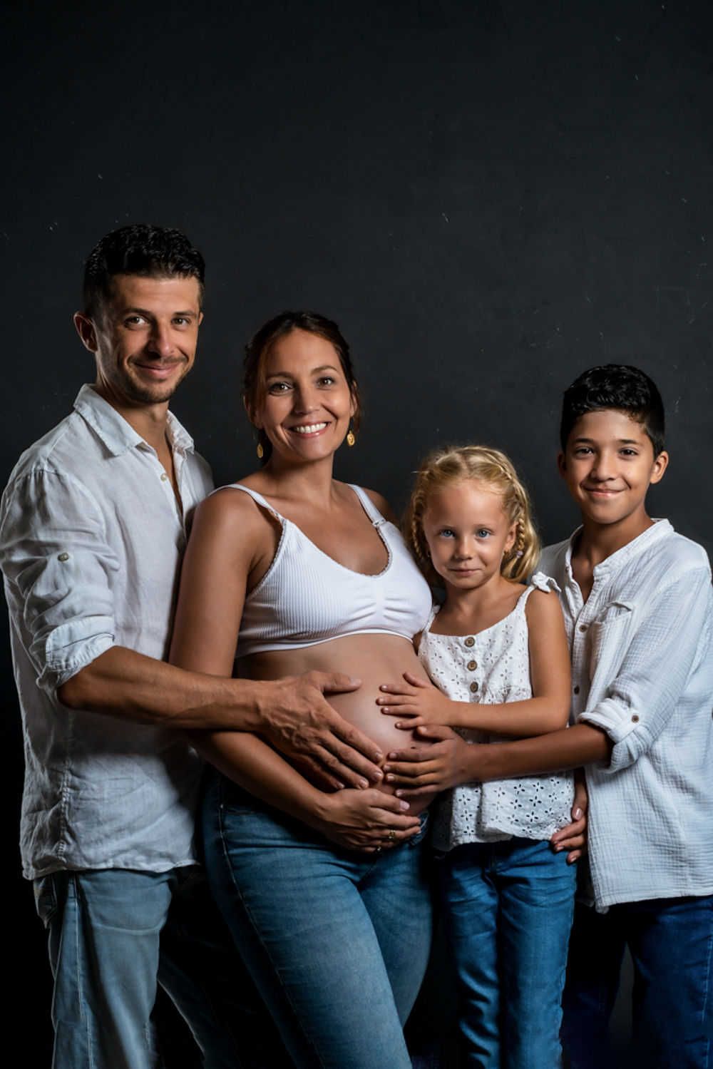 Une femme enceinte en haut blanc et jean pose avec sa famille sur un fond sombre pour une photo de maternité.