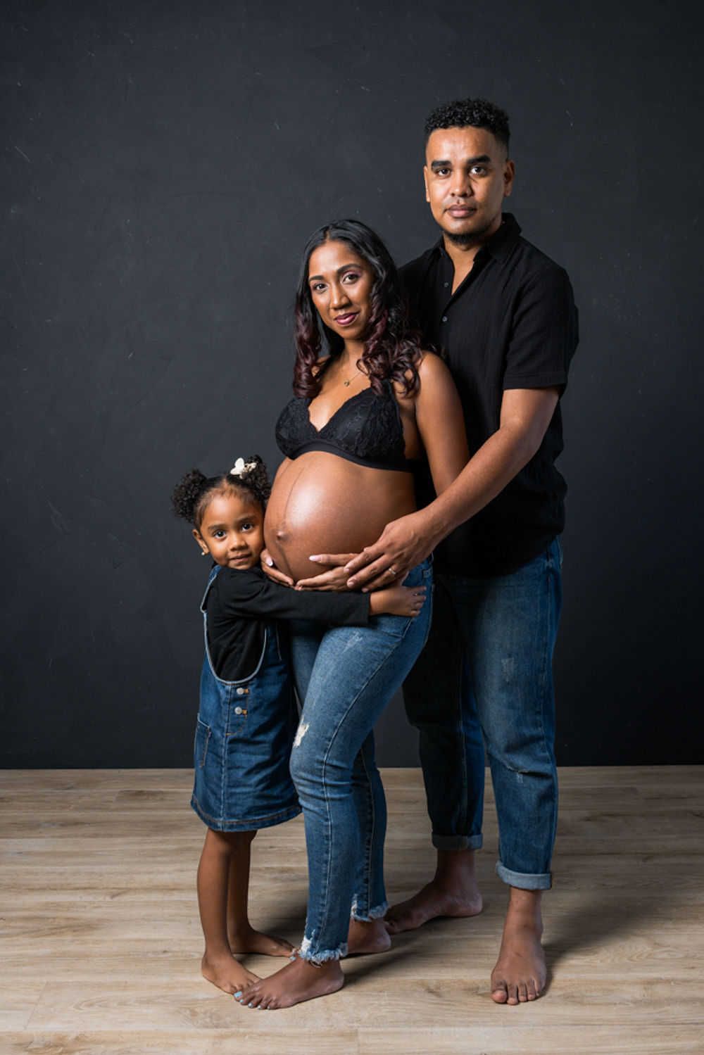 Une photo de famille de maternité sur fond sombre avec des poses pieds nus sur un parquet.