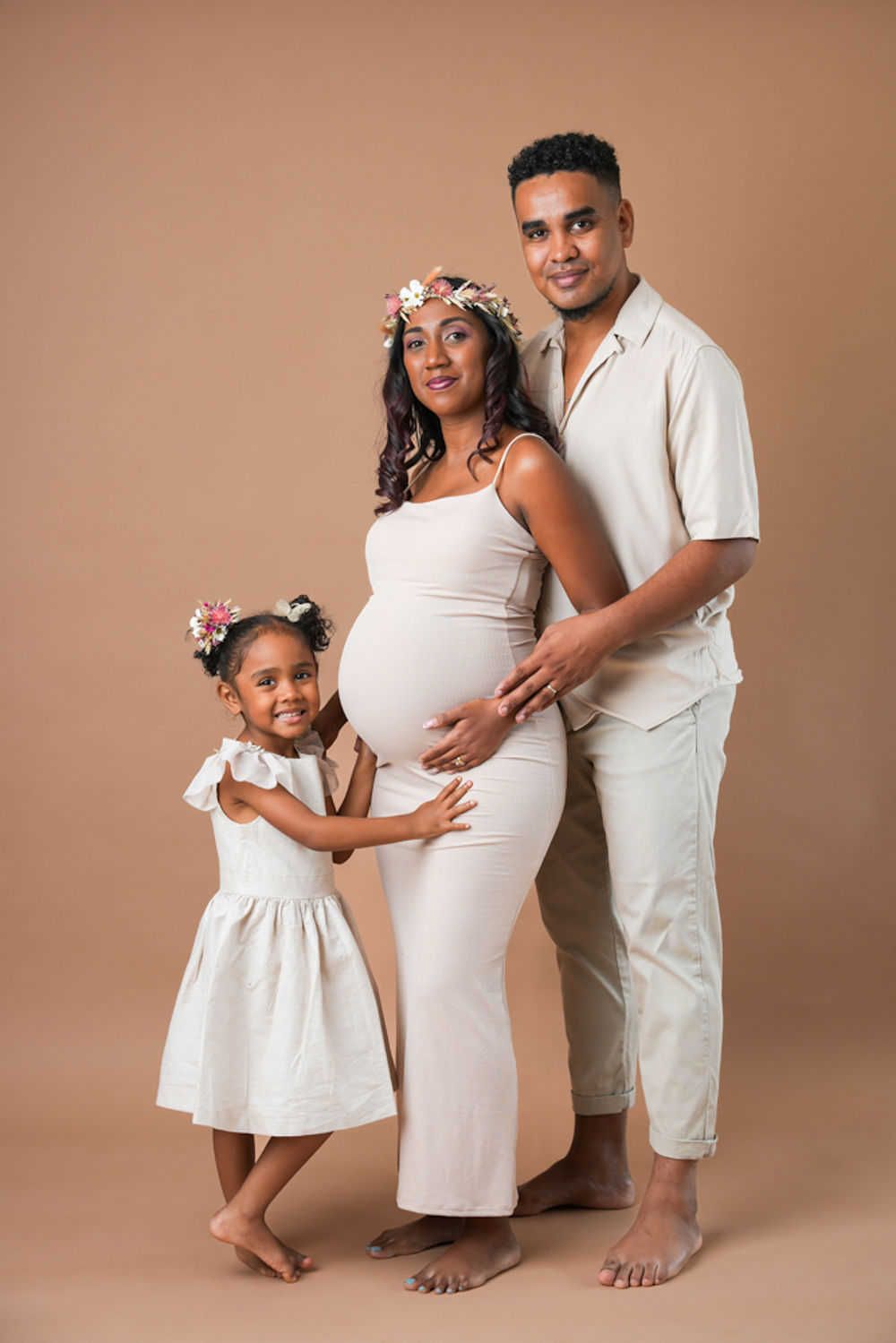 Famille posant ensemble pour une photo de grossesse dans des tenues blanches assorties sur fond beige avec des accents floraux.