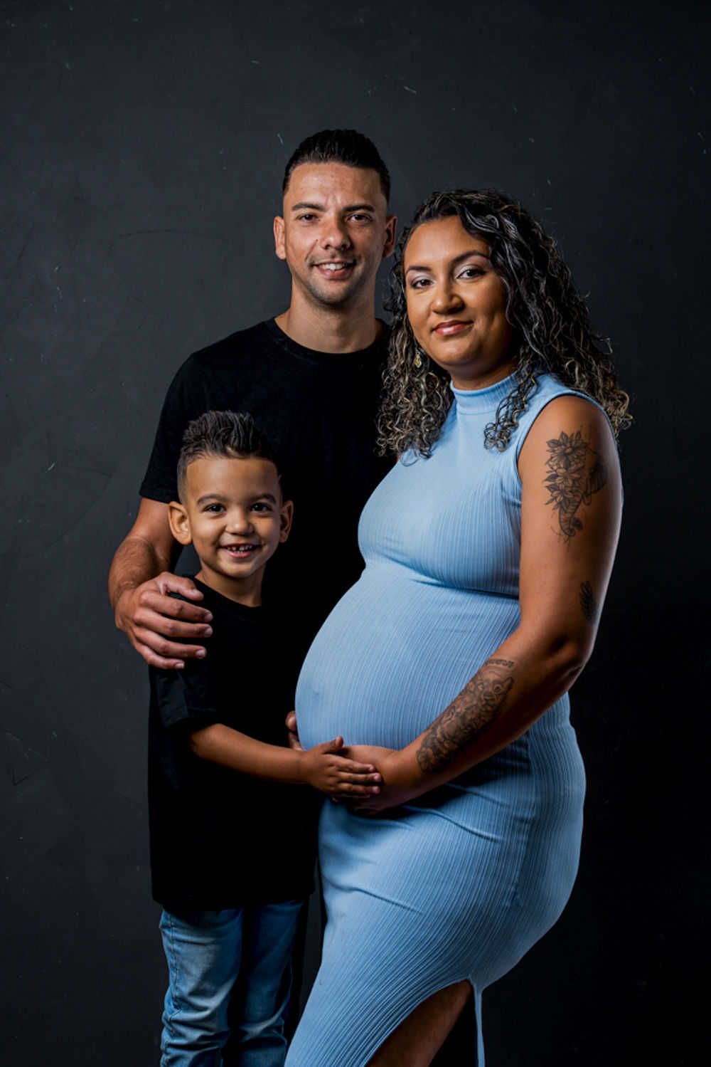 Un portrait de maternité familial avec une femme enceinte en robe bleue posant avec ses proches sur fond sombre.