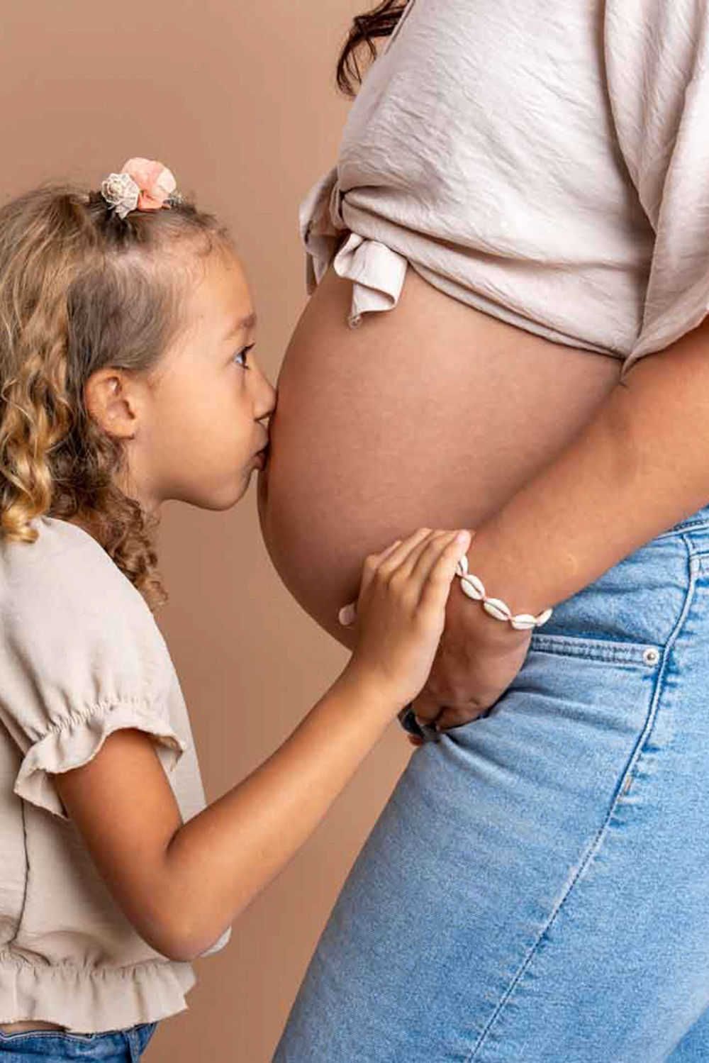 Un enfant vêtu d'un haut beige touche un ventre de femme enceinte portant un jean sur un fond neutre.