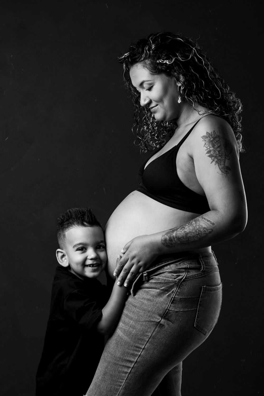 Portrait en noir et blanc d'une future maman aux cheveux bouclés et aux tatouages ​​partageant un moment de joie avec un enfant souriant.