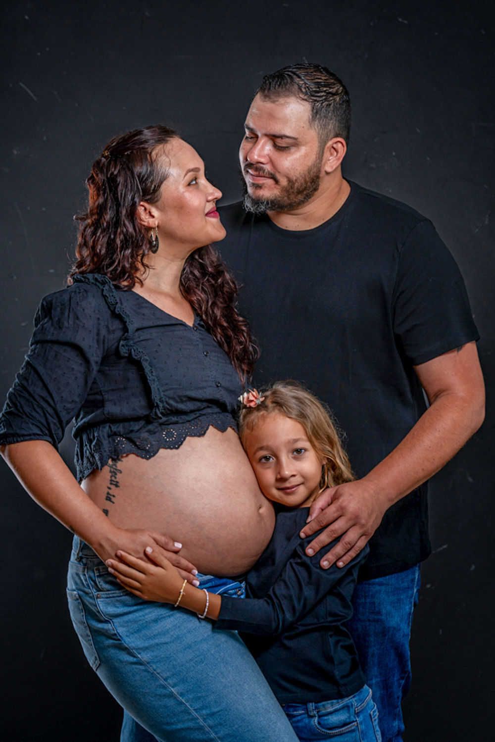 Une famille pose ensemble sur un fond sombre tout en célébrant une grossesse à venir dans des tenues bleu marine assorties.