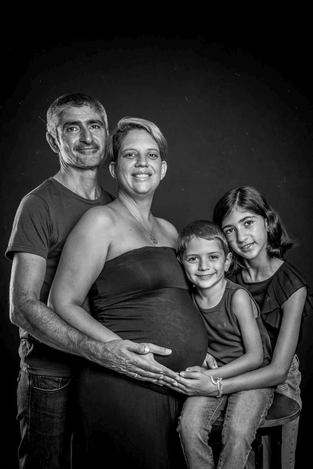Portrait de famille en noir et blanc montrant de futurs parents et deux jeunes enfants posant ensemble sur un fond sombre.