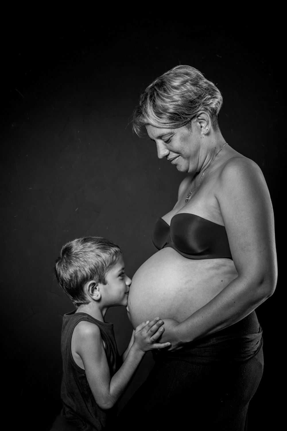 Un portrait en noir et blanc capture un moment intime entre une femme enceinte et un jeune enfant sur un fond sombre.