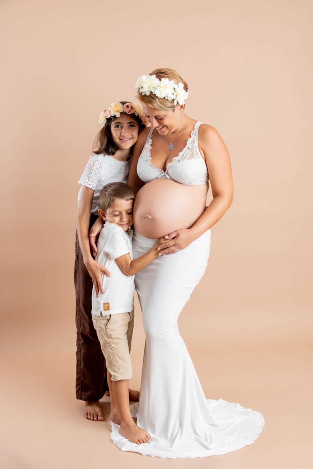 Portrait de maternité avec robe en dentelle blanche et couronnes de fleurs sur fond beige avec des enfants.