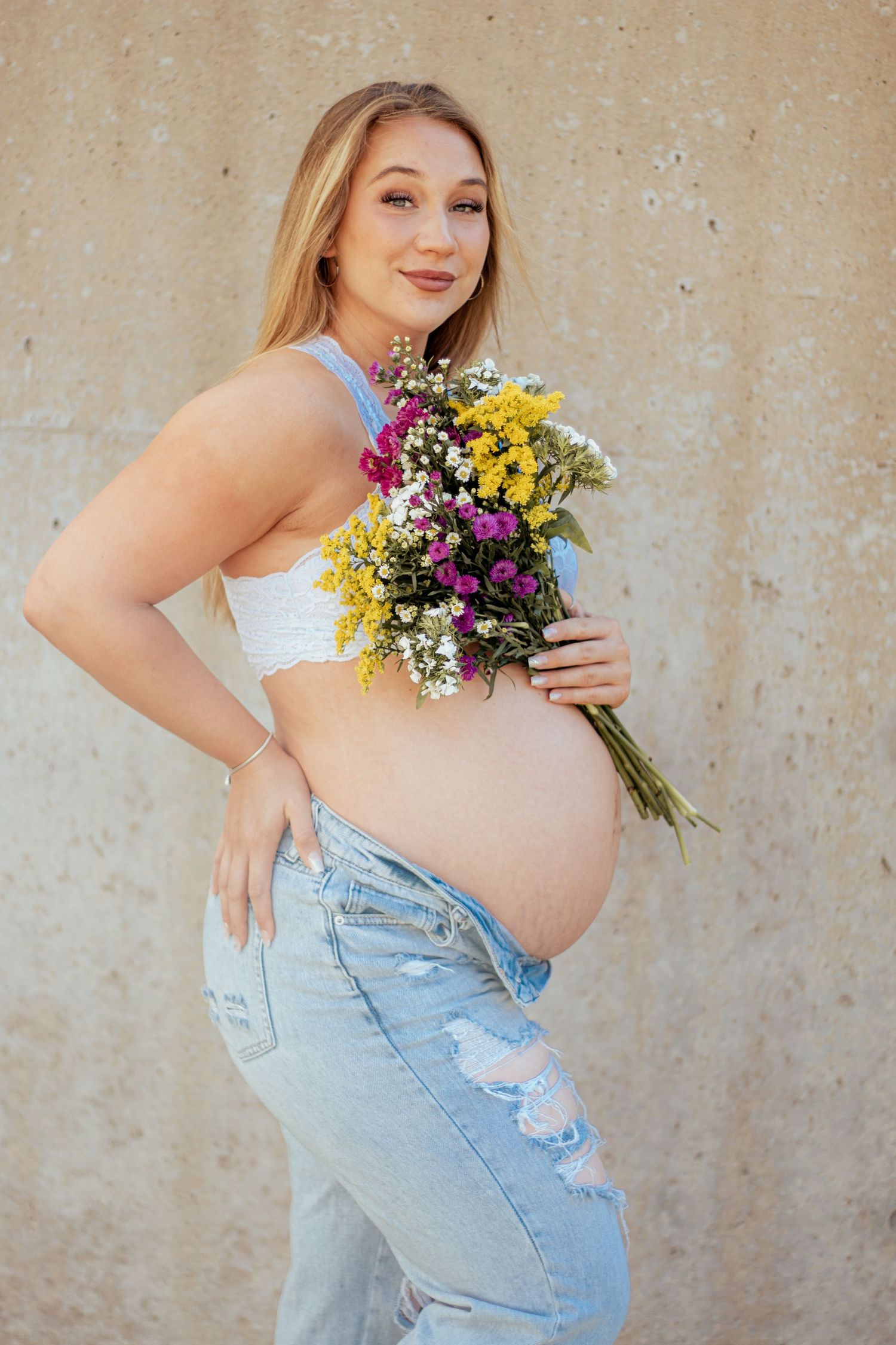 Expecting mother in ripped jeans and crop top holds colorful wildflower bouquet against beige wall.