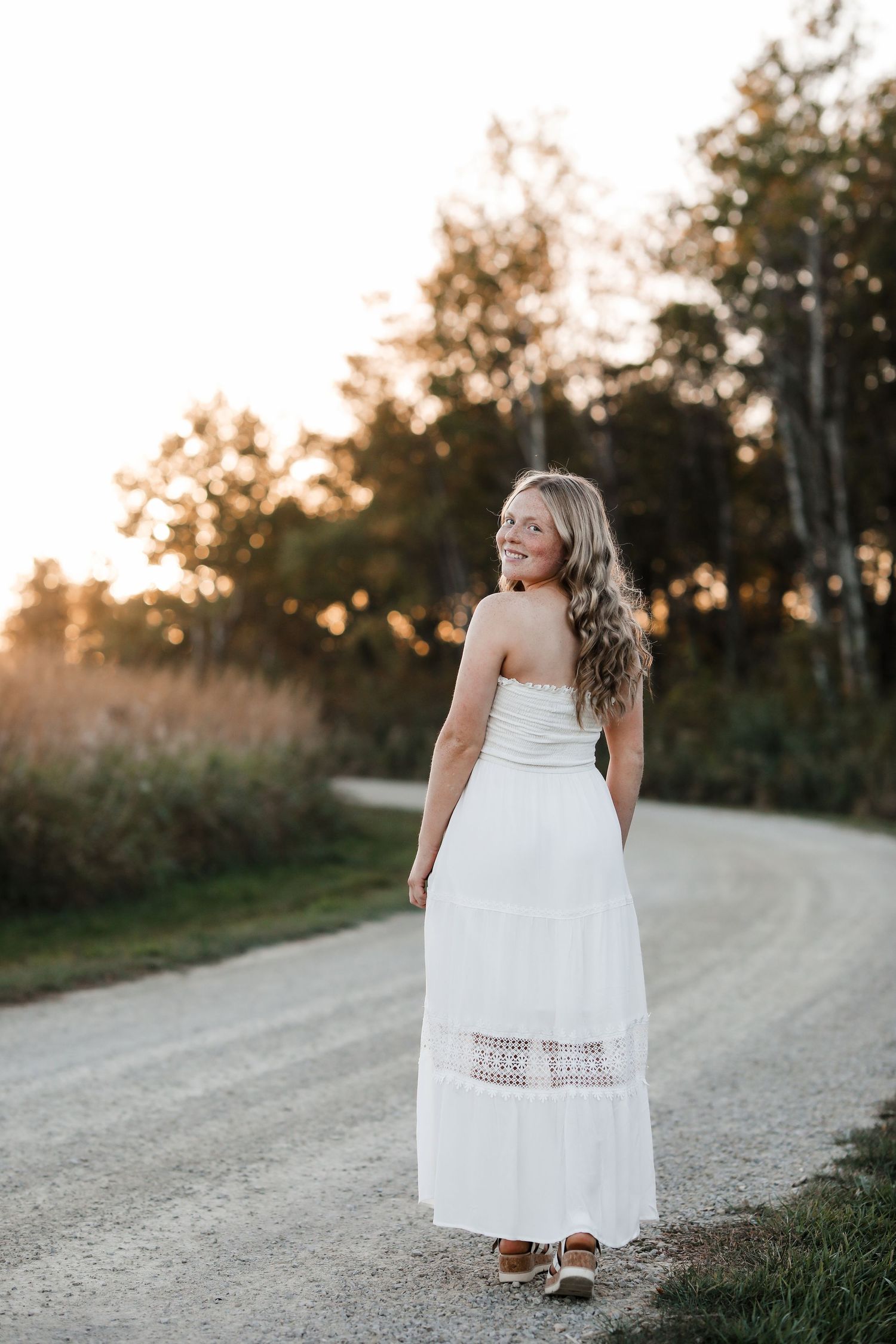 Smiling blonde girl in white strapless maxi dress walking on gravel road at sunset.