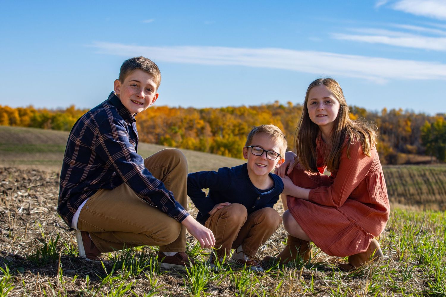 Three siblings sit together on a grassy hill during autumn with fall foliage in the background.
