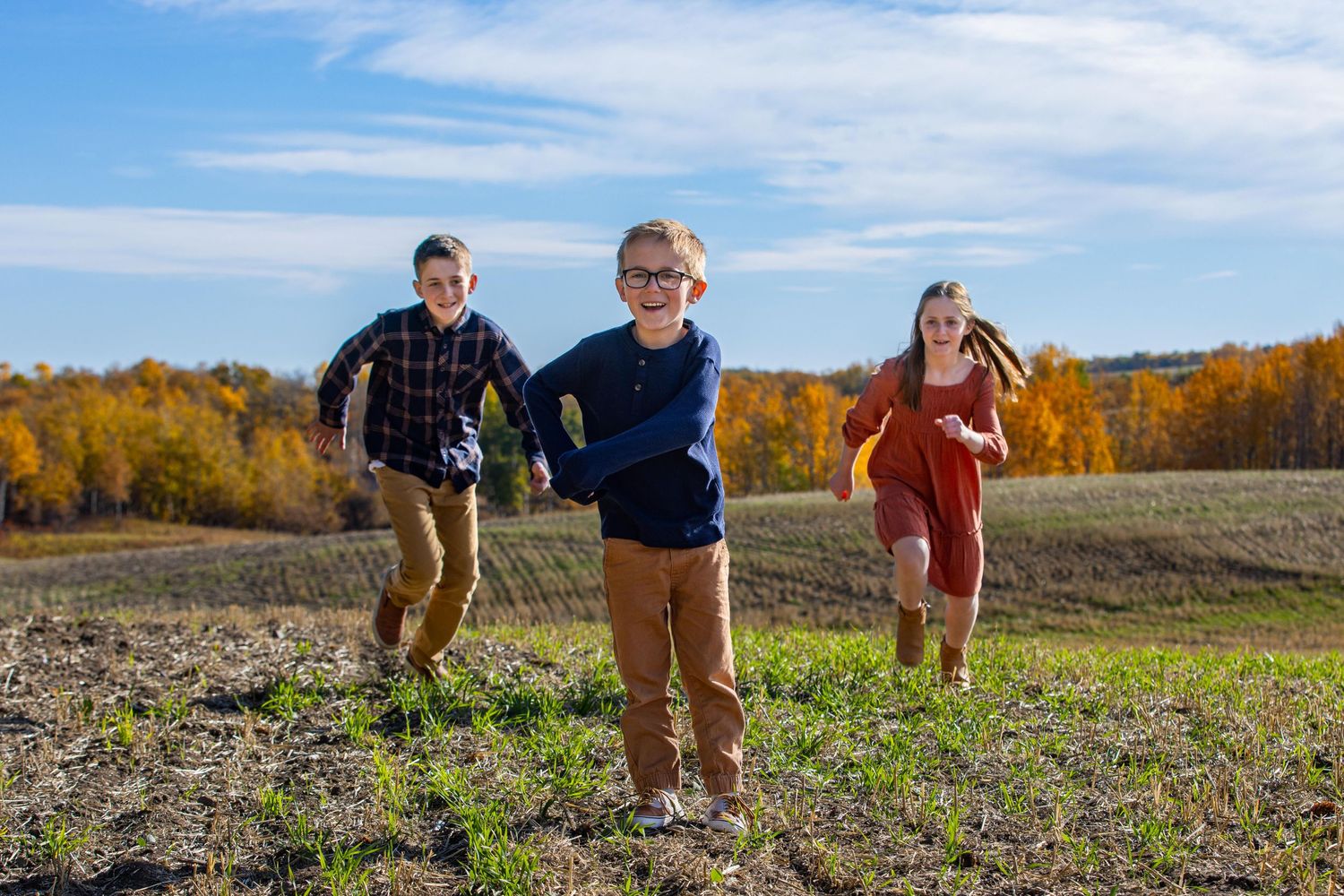 Three siblings run and play together in a field with colorful autumn trees in the distance.