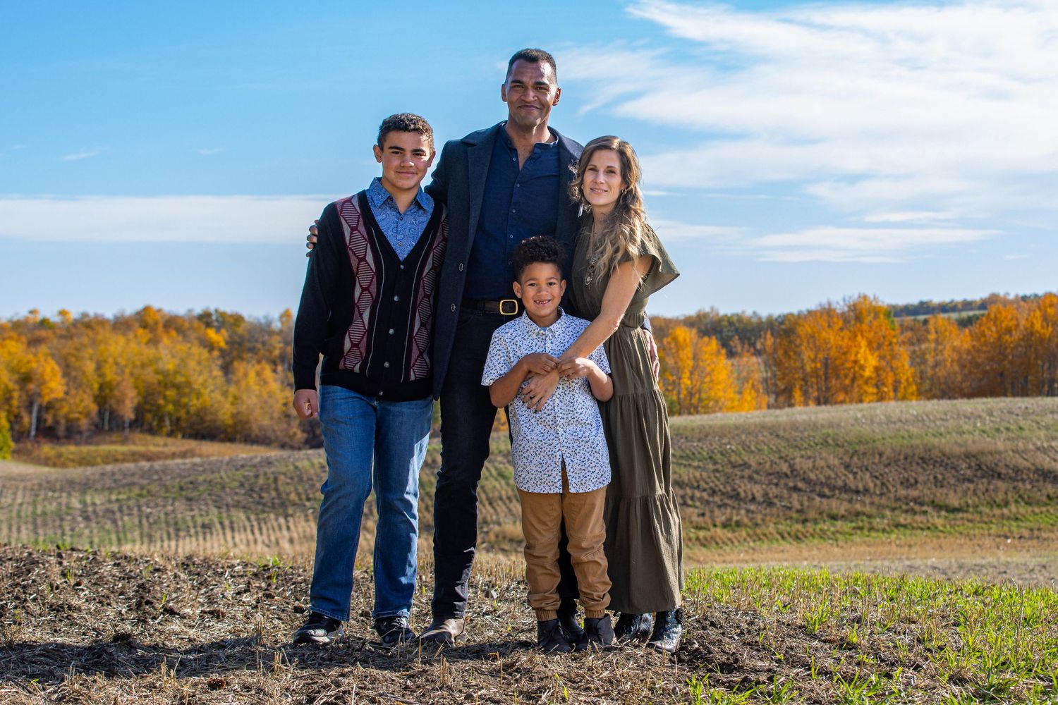 A family stands together on a hillside during autumn with golden trees and blue sky in the background.
