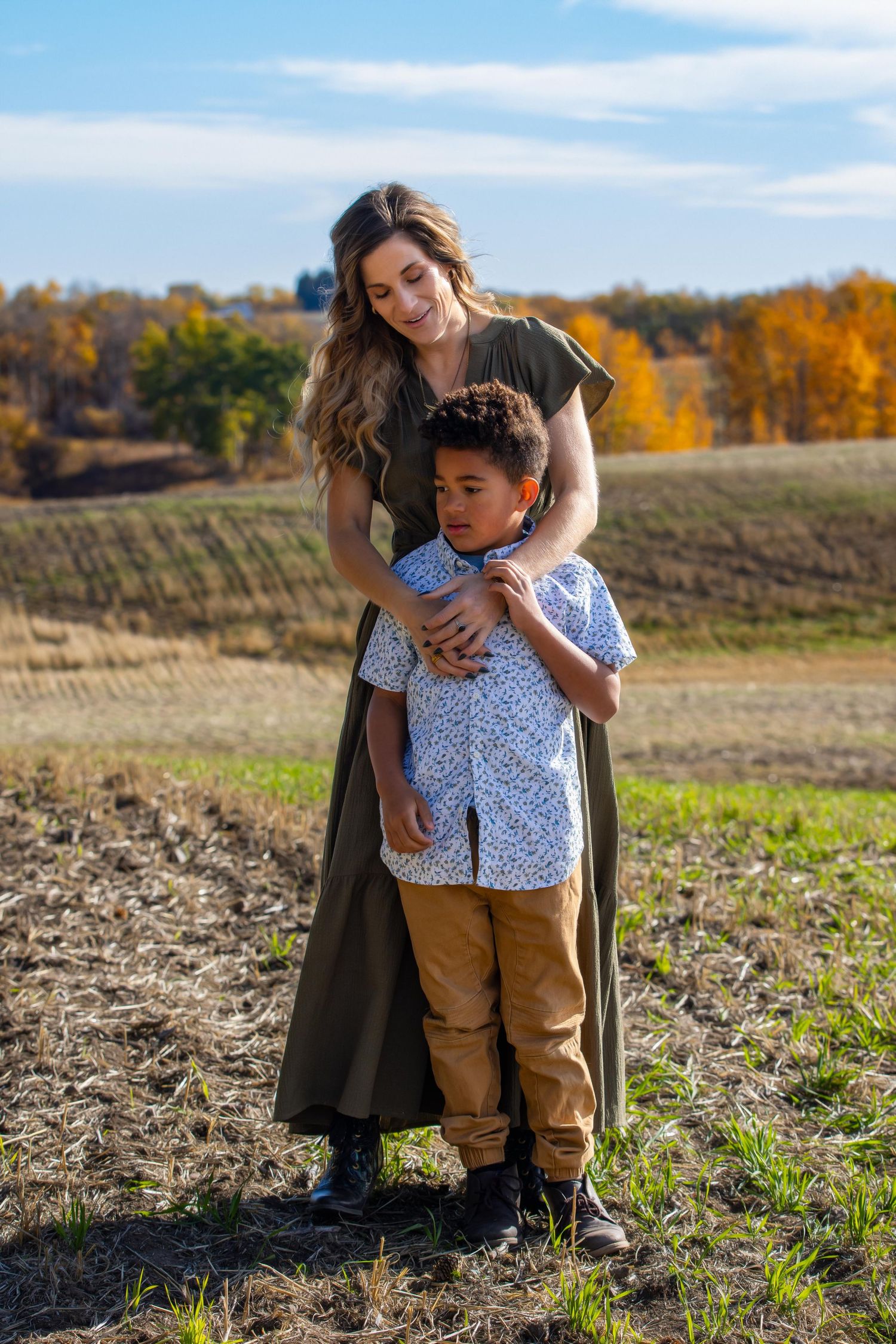 A tender family moment captured in a field during autumn with fall foliage and a blue sky in the background.