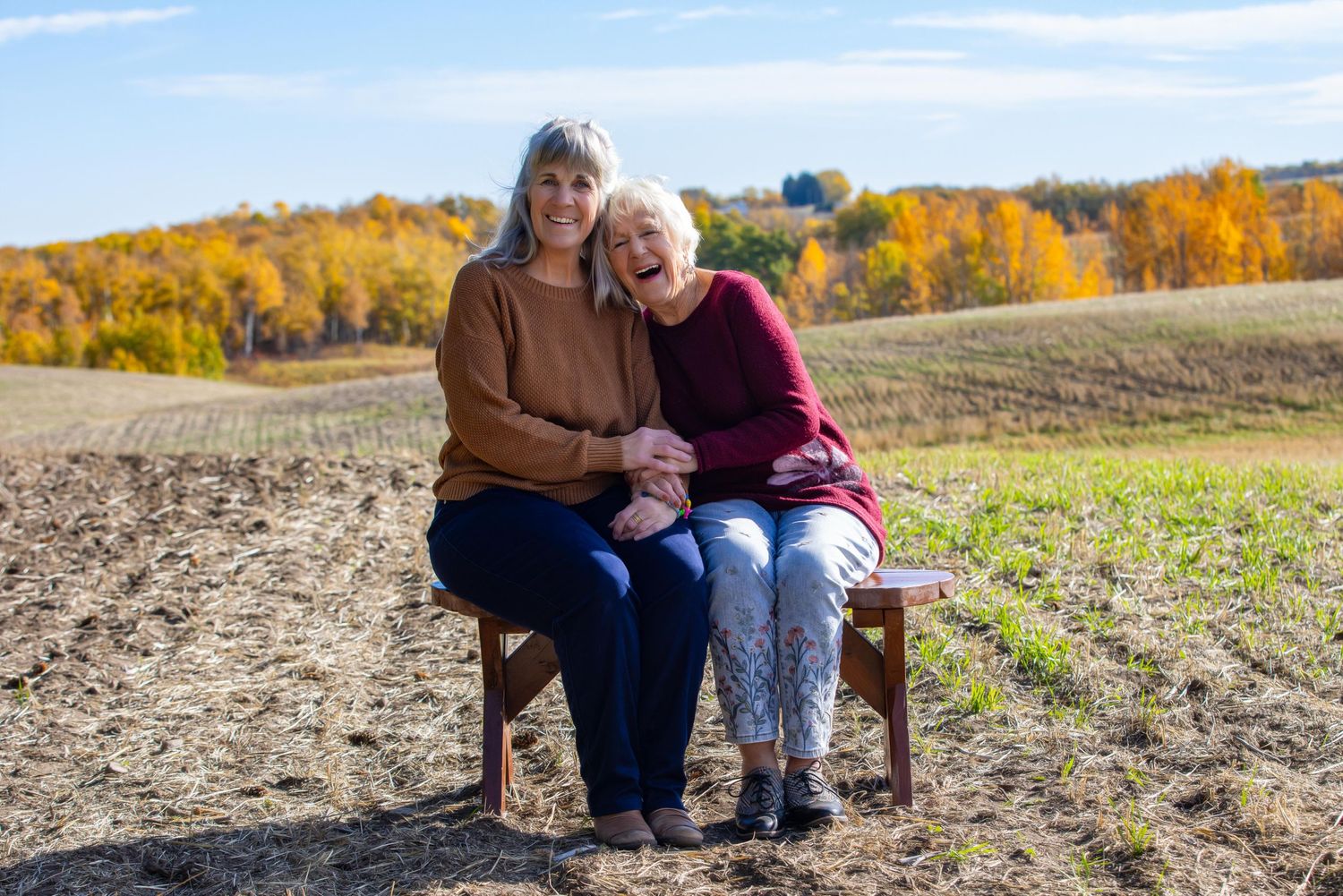 Two people sit close together on a wooden bench in a field during autumn with golden trees in the background.