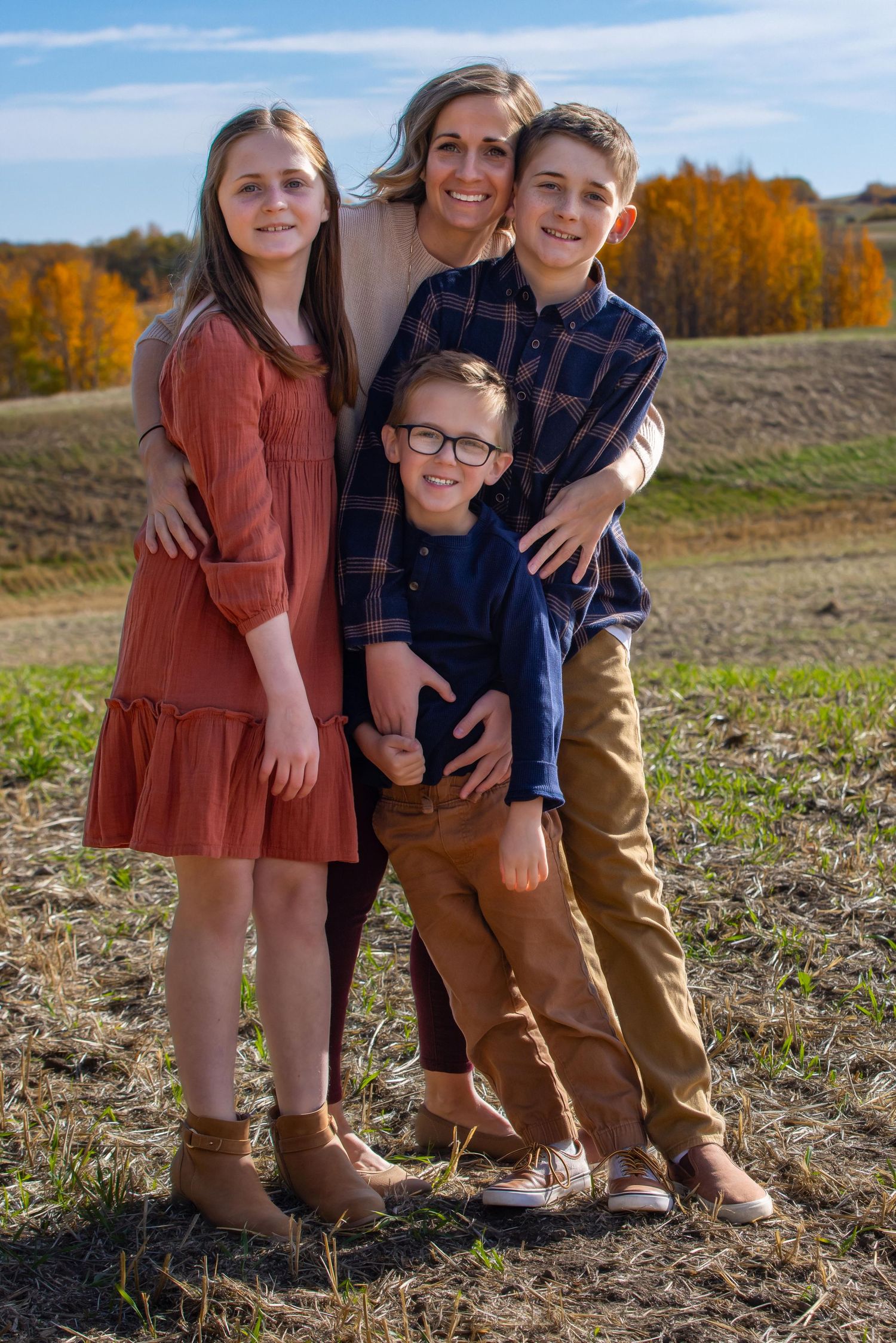 A group of four people pose together outdoors against a backdrop of fall foliage and blue sky.
