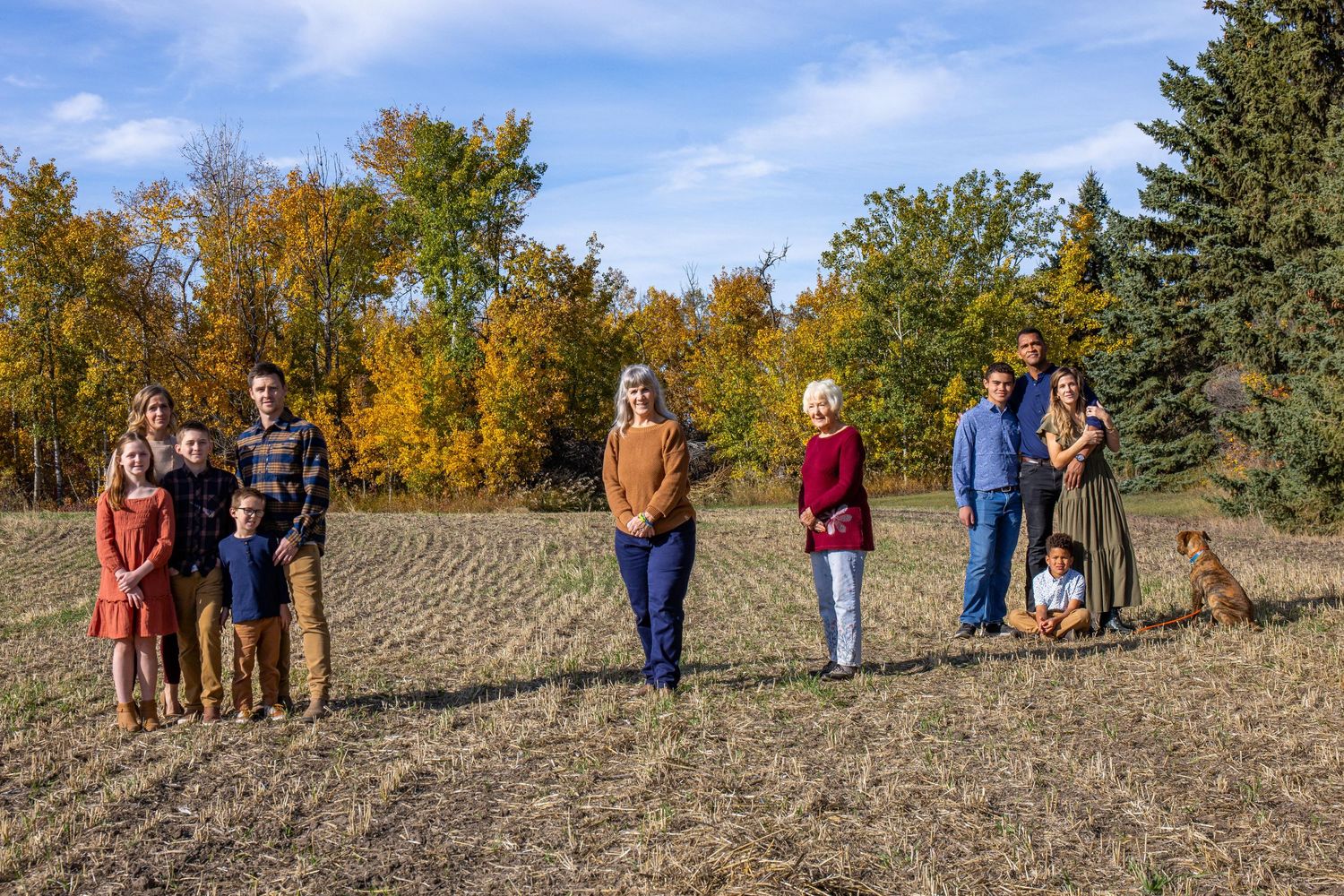 Group of people standing together in a harvested field on a sunny autumn day with golden trees in the background.