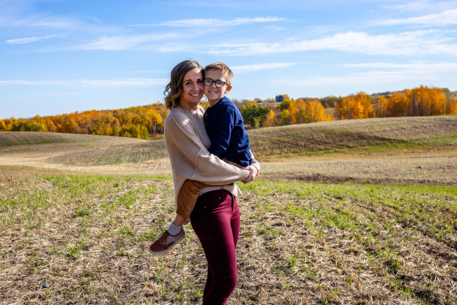 A couple share a playful moment in an autumn field with golden trees in the background on a sunny day.