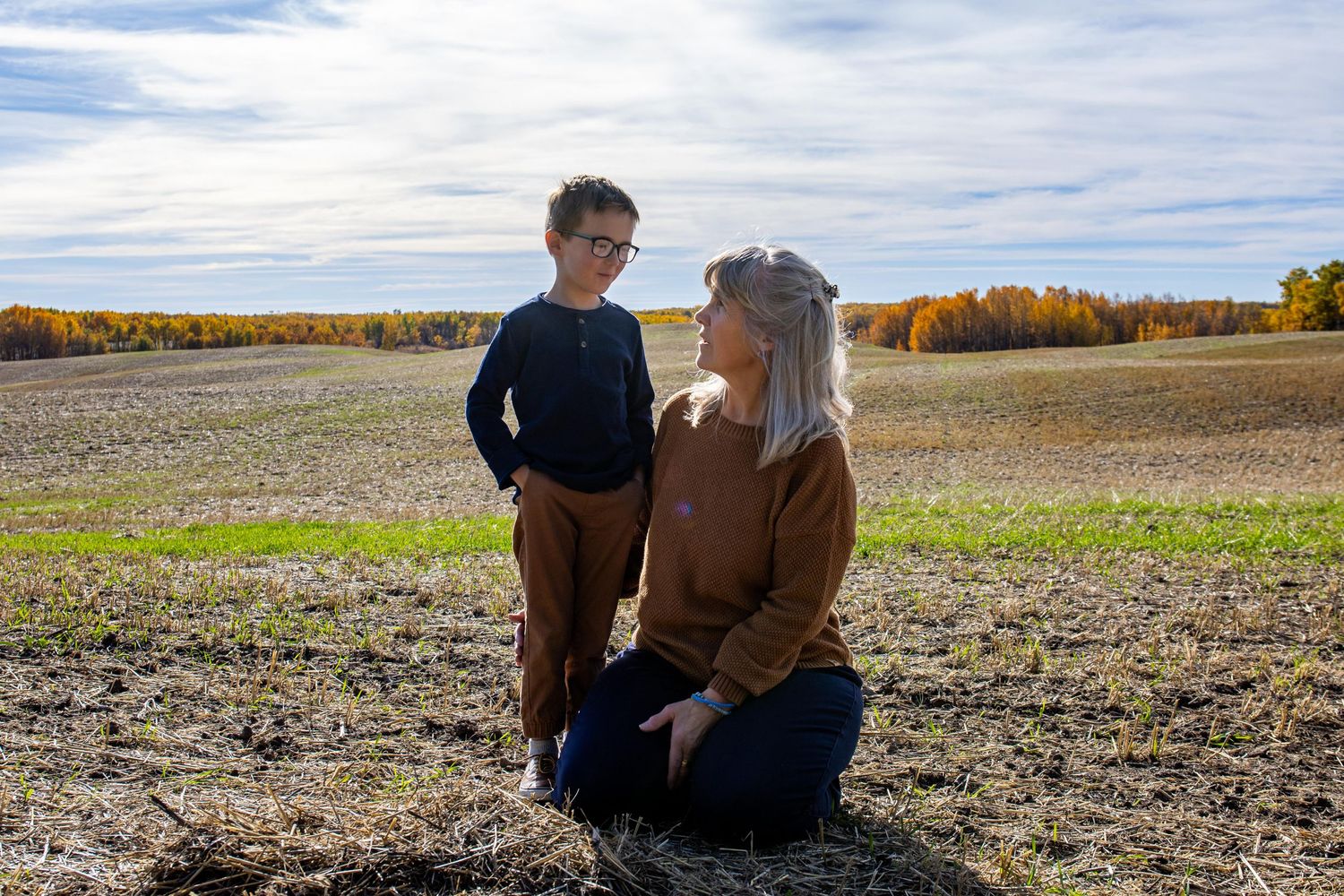 Two people pose together in a harvested farm field on a sunny autumn day with colorful fall foliage in the background.
