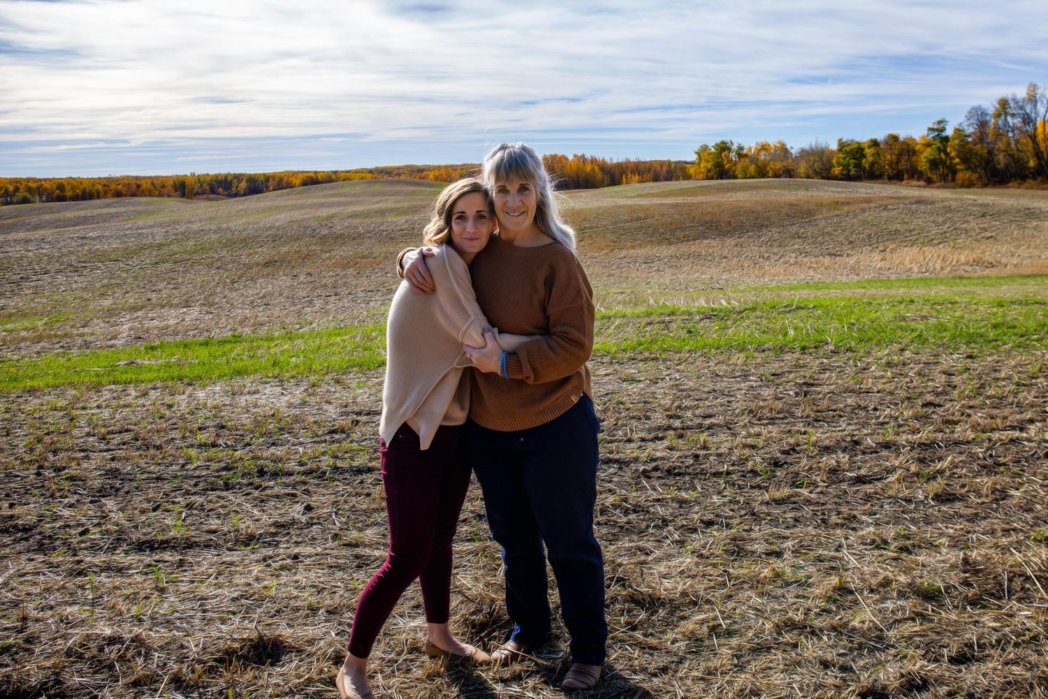 Two people embrace while standing in a harvested field surrounded by fall foliage on a bright afternoon.