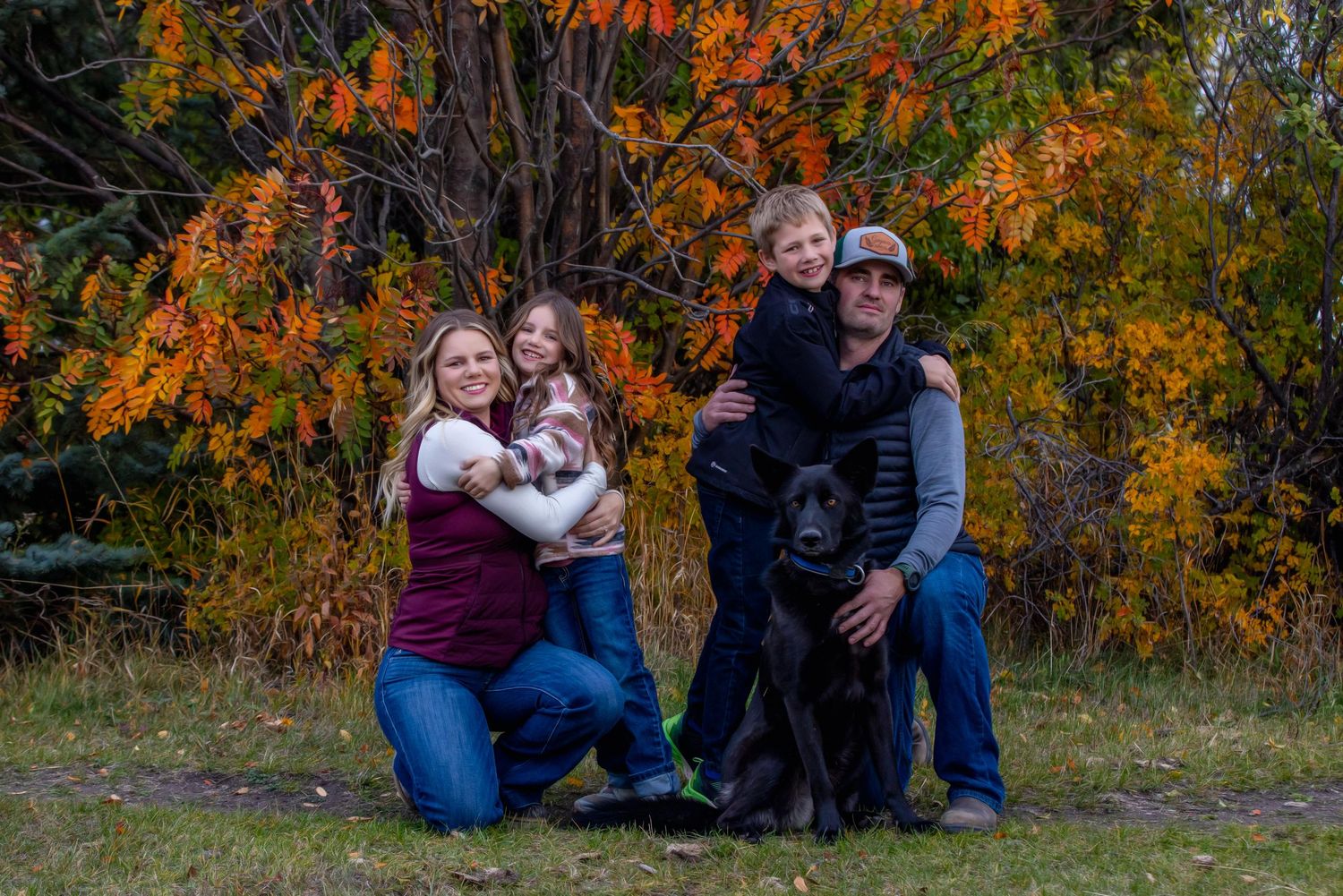A family group poses together outdoors against vibrant orange autumn foliage with their black dog in the foreground.