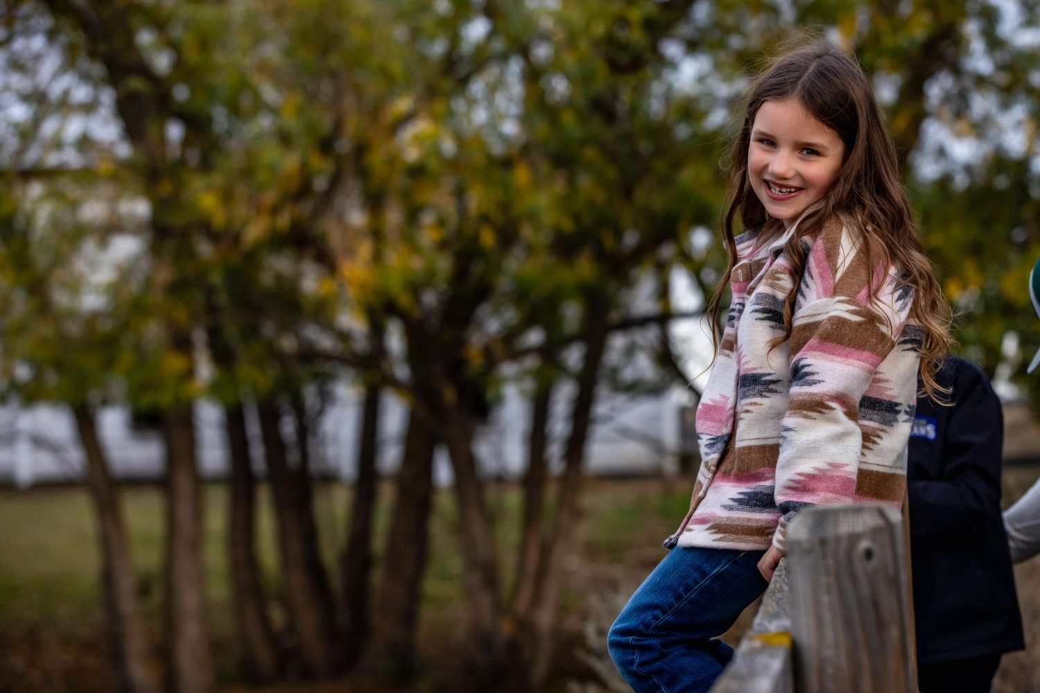 A young person in a plaid sweater sits outdoors smiling against a blurred autumn background.