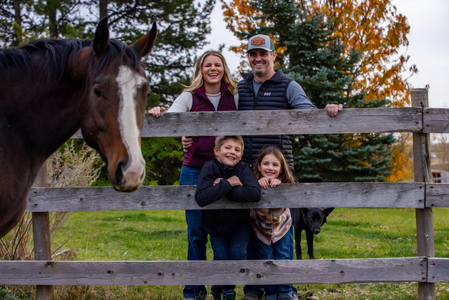 Family and horse pose at wooden fence on autumn day with colorful fall foliage in background.