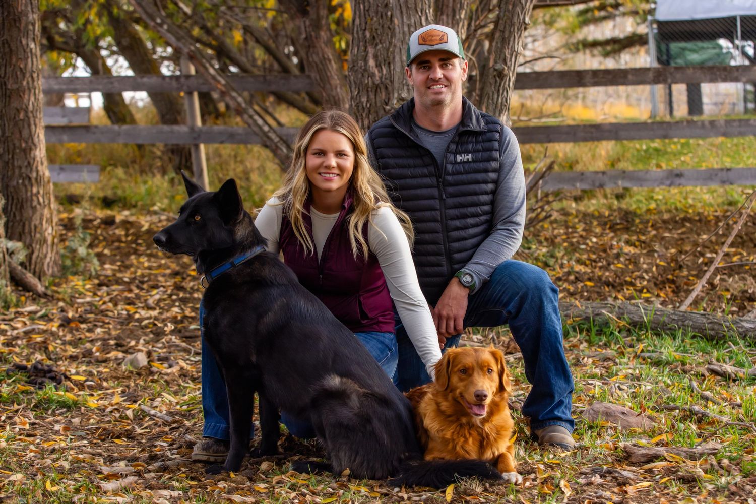Two people sit outdoors with their black Labrador and golden-haired dog on autumn leaves with a fence in the background.