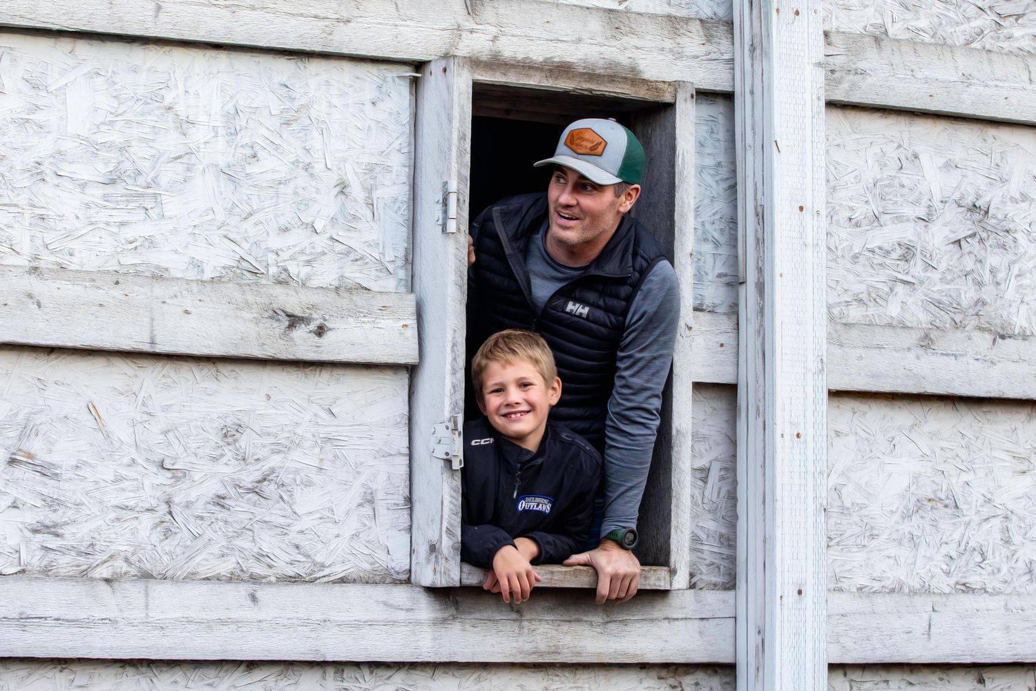 Two people peek out and smile from a rustic wooden window frame of a weathered white building.
