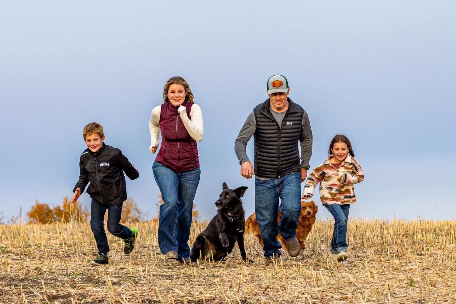 A group walks together with their black dog across a golden hilltop field against a blue sky background.