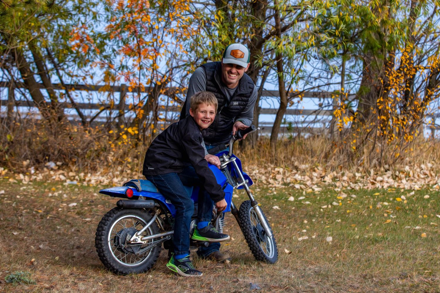 Two people sitting on a blue dirt bike on an autumn trail with fallen leaves and wooden fence in background.