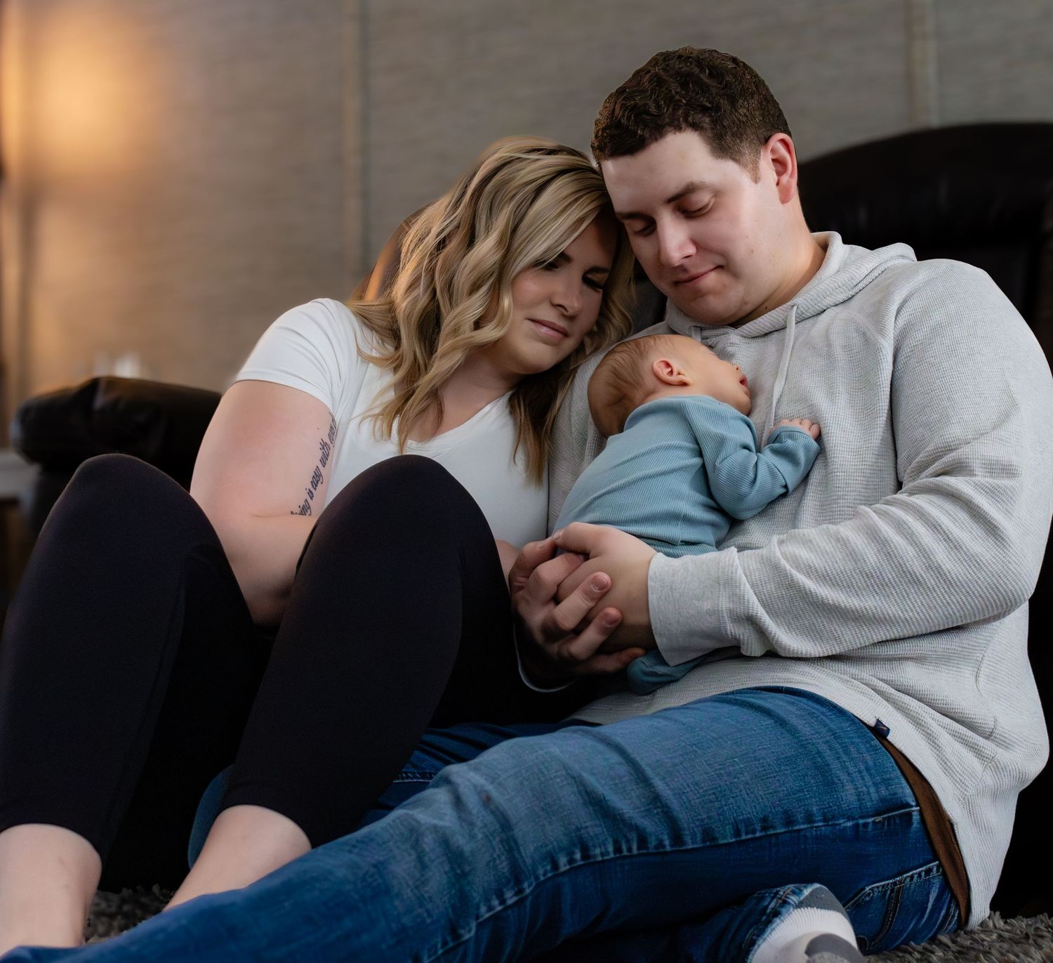 A couple sits close together on a couch, sharing a tender moment with a sleeping baby in a peaceful home setting.