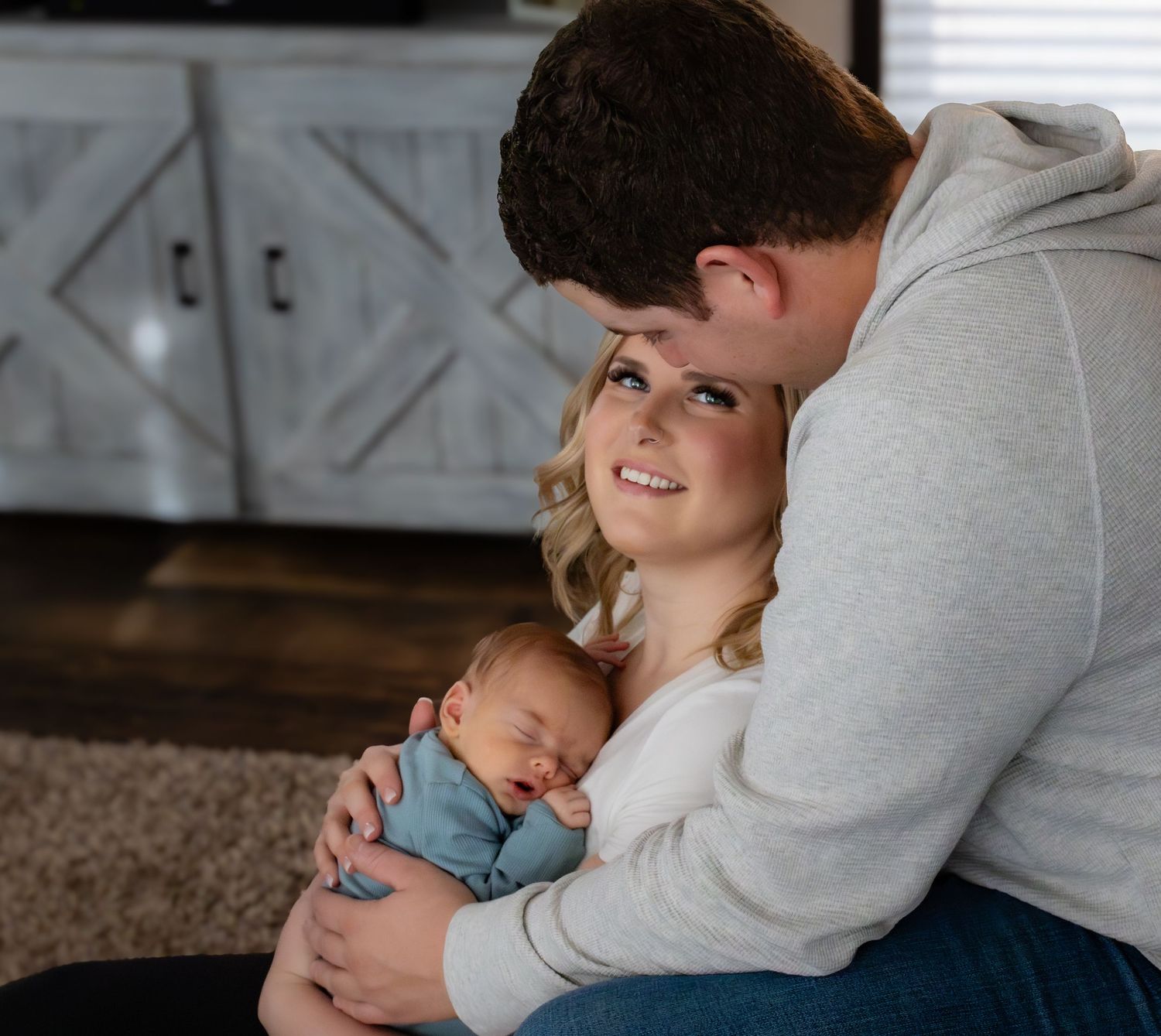 A couple tenderly embracing their sleeping newborn baby while sitting on carpeted floor at home.
