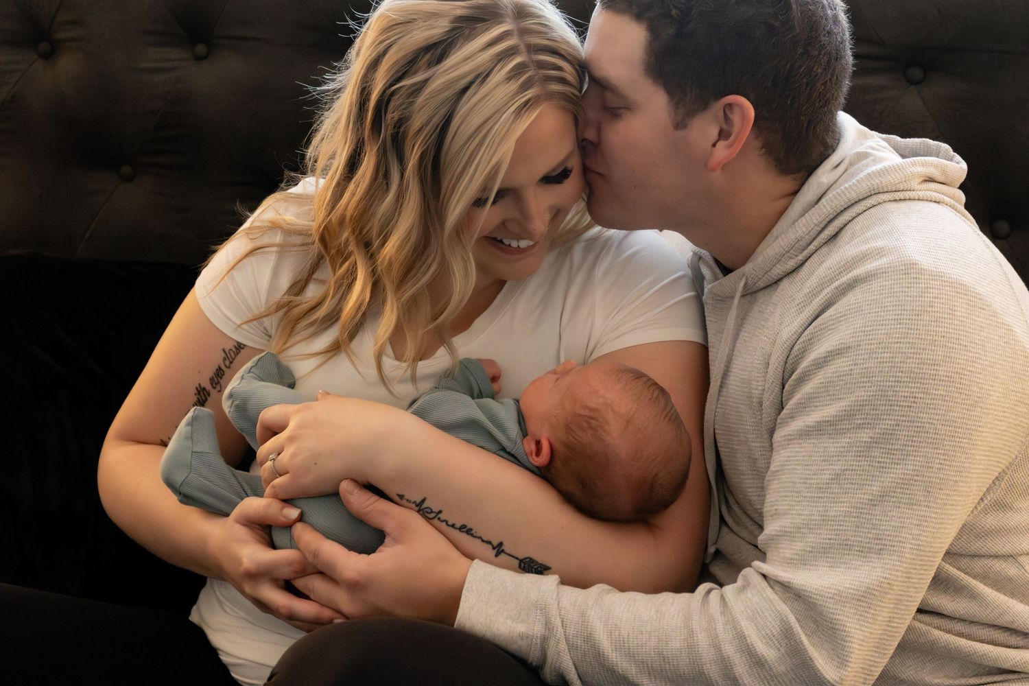 A couple cuddles together on a dark couch with a sleeping newborn baby wearing gray clothing.