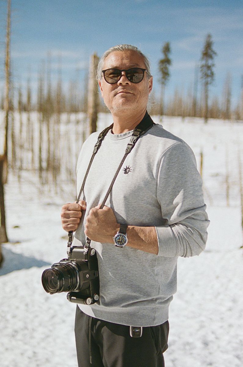 A photographer in a grey sweater holding a vintage camera against a snowy winter landscape.