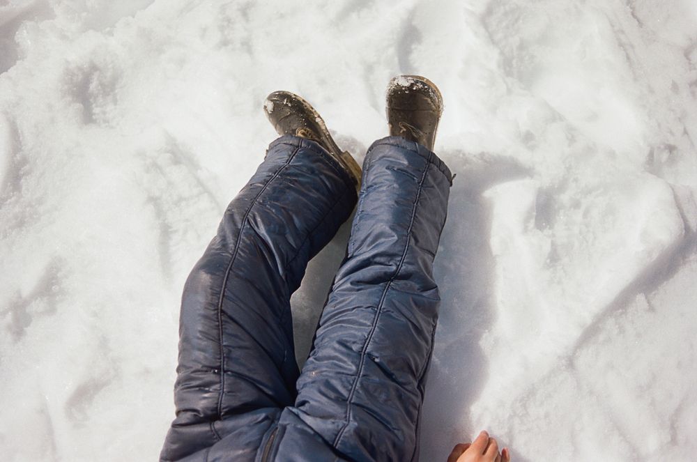 A pair of snow-covered legs wearing dark pants and boots viewed from above on white snowy ground.