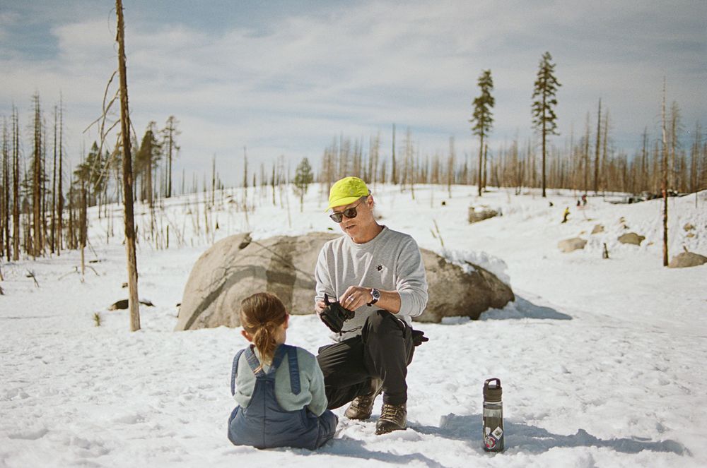 Two people taking a break in the snow with a water bottle near bare winter trees.
