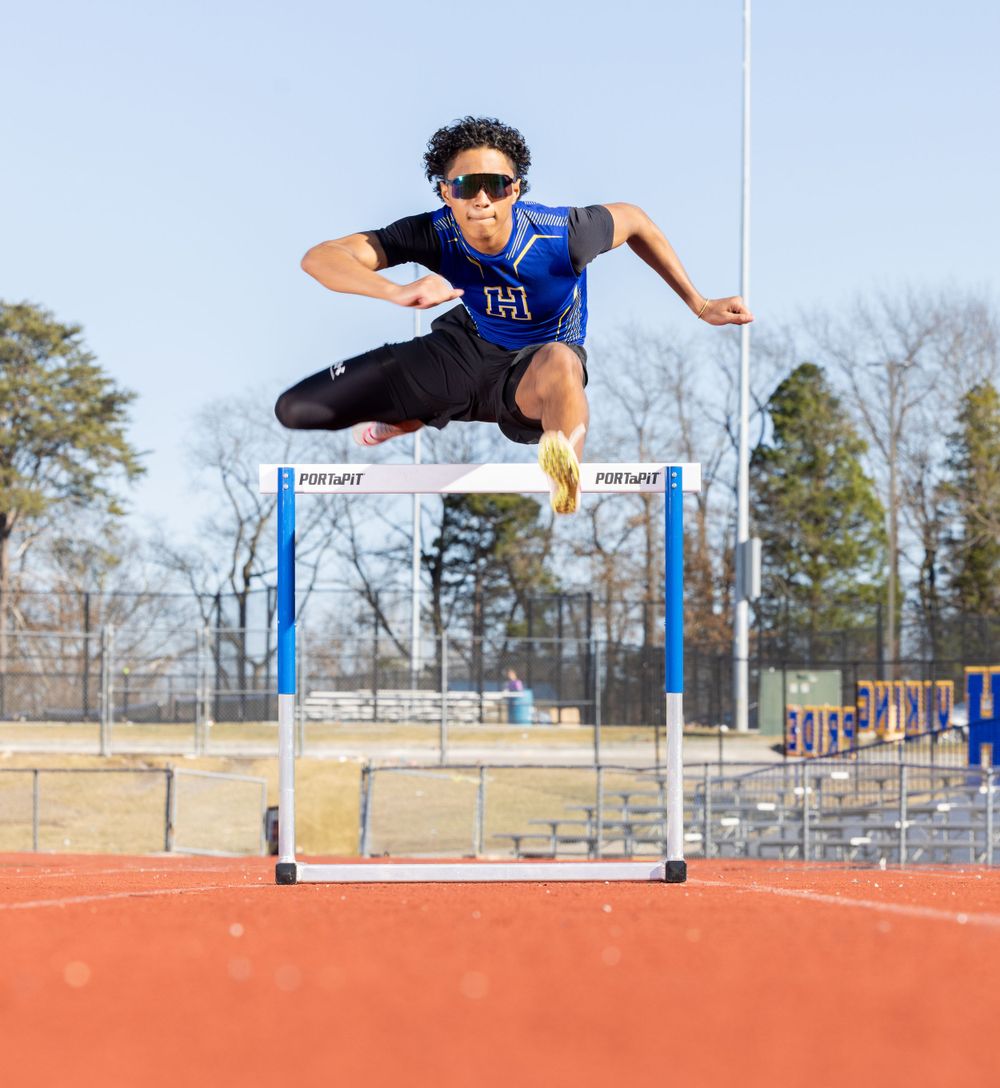 Track athlete leaping over blue hurdle on red running track during outdoor competition.