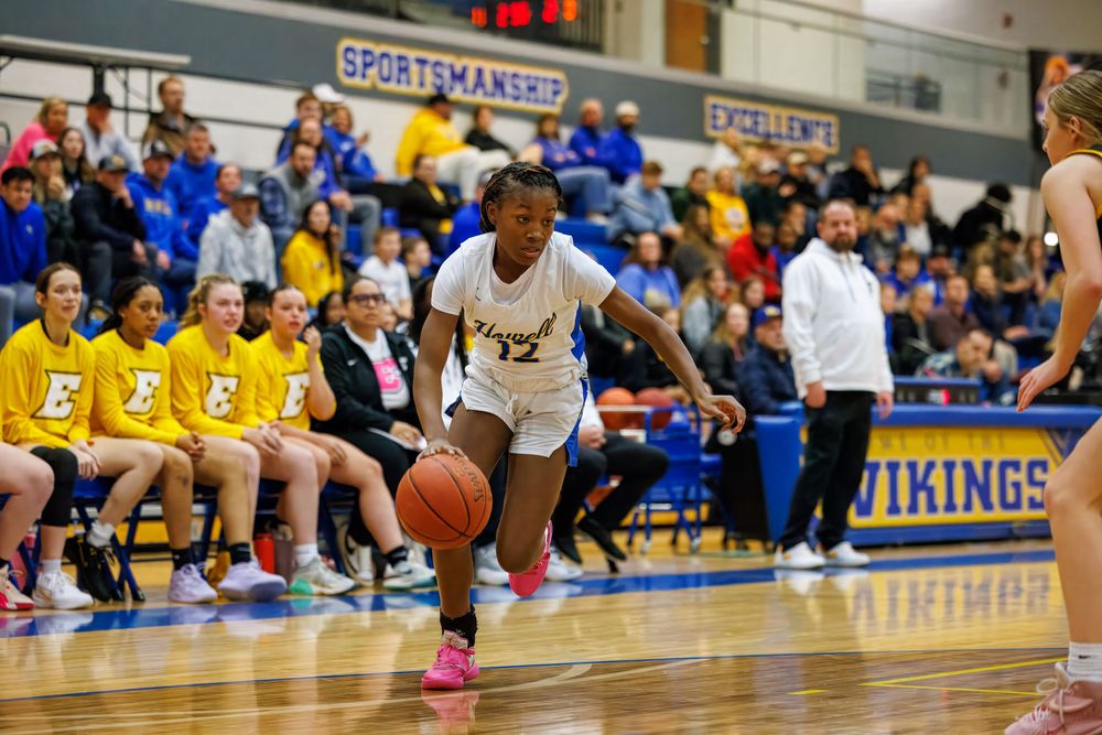 Basketball player in white jersey dribbles the ball during intense game action on wooden court with spectators in background.