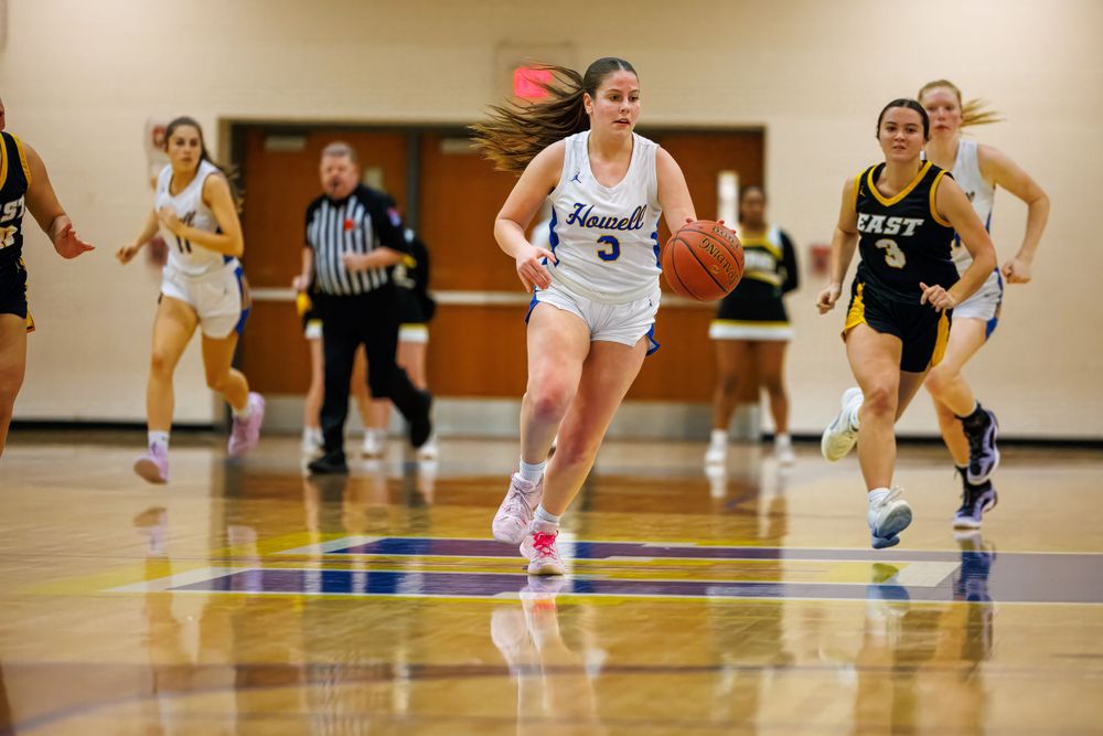Basketball player drives down court in white uniform while opponents in black jerseys give chase during indoor game.