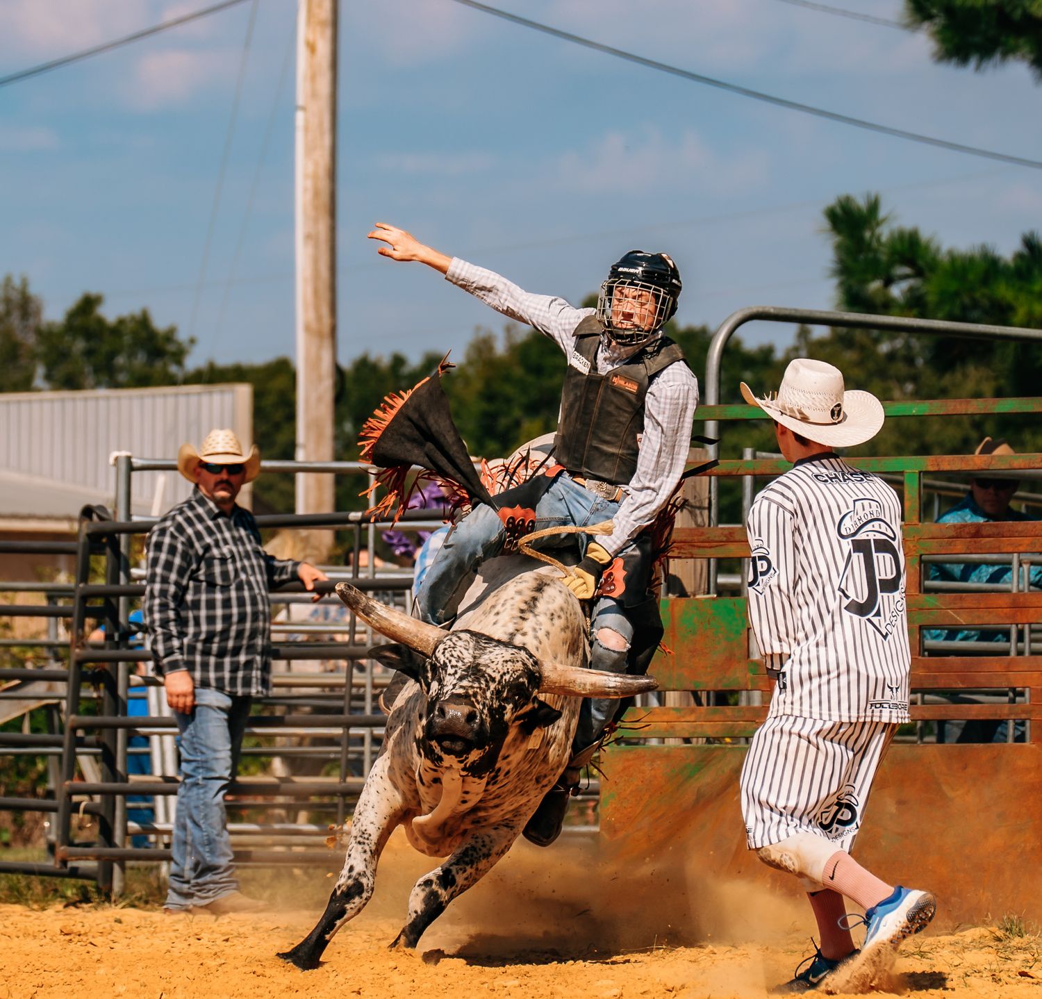 Bull rider competing on bucking bull while rodeo clowns stand ready in arena.