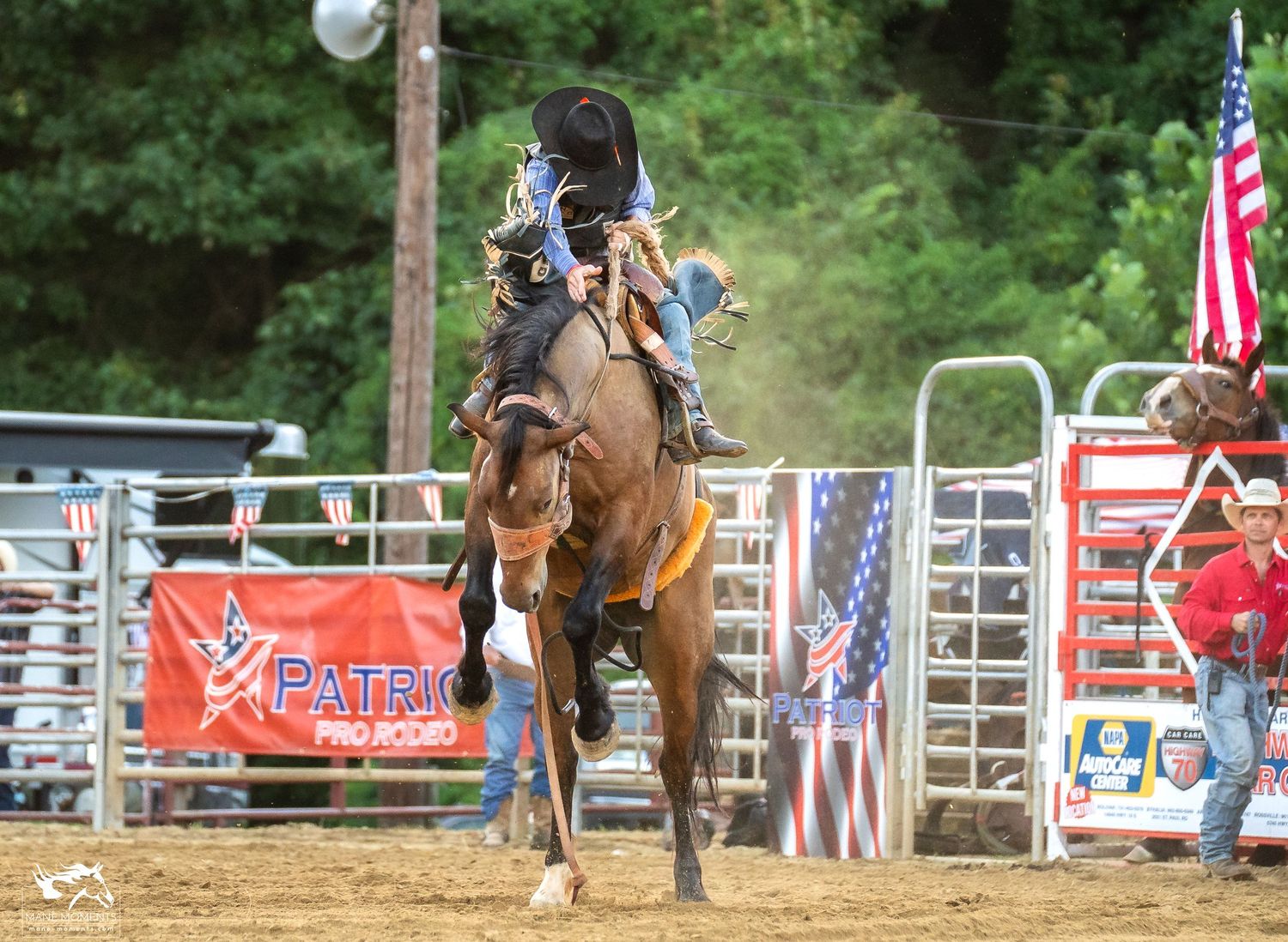 A bronc rider competes in a rodeo arena with American flags visible in the background.
