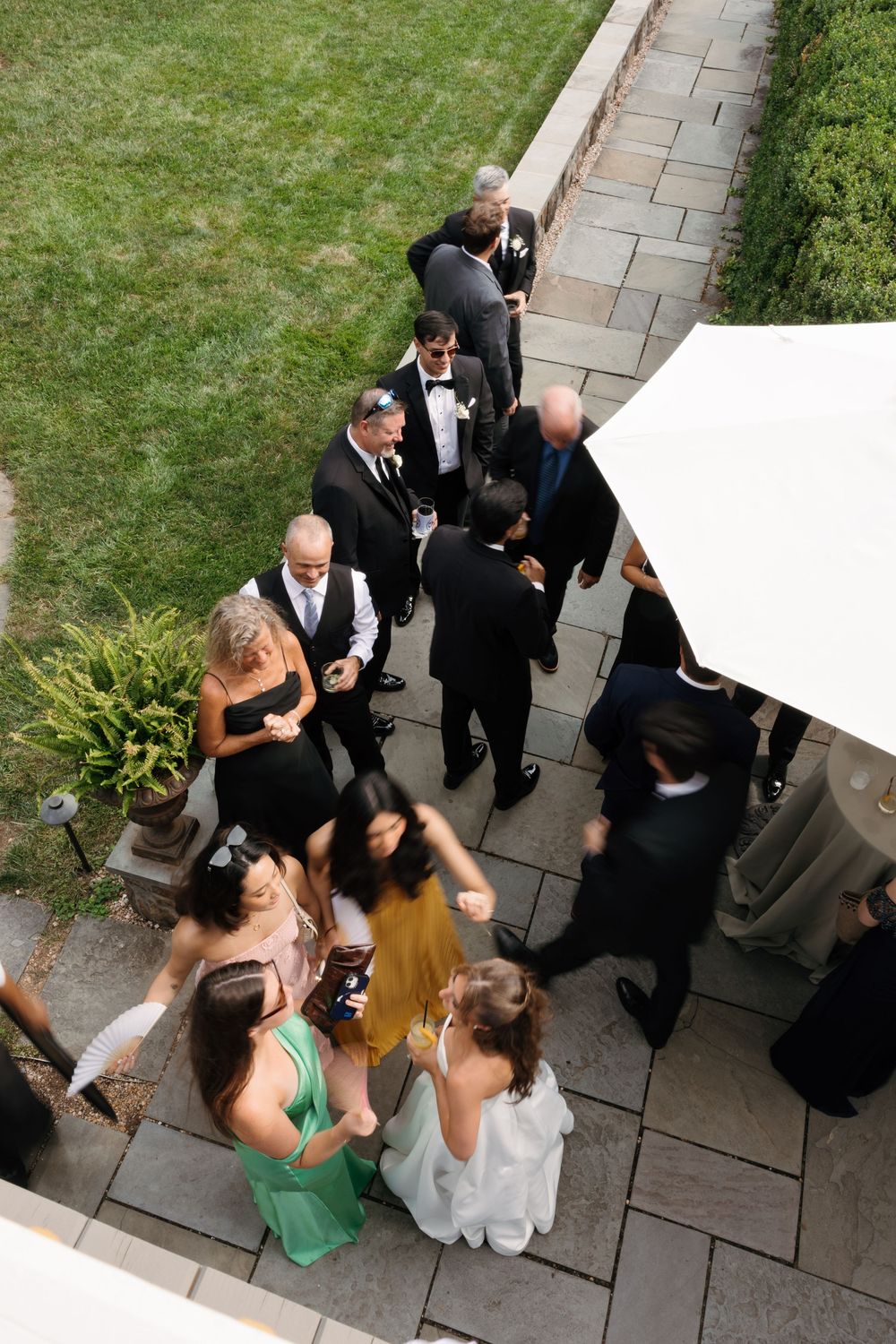 Aerial view of elegantly dressed wedding guests mingling on a stone patio surrounded by lush green lawn.