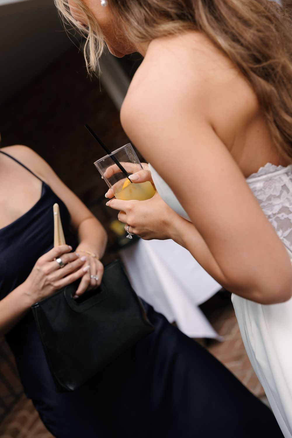Two women at a wedding, one in white lace holding a champagne glass, the other in navy blue holding a clutch.