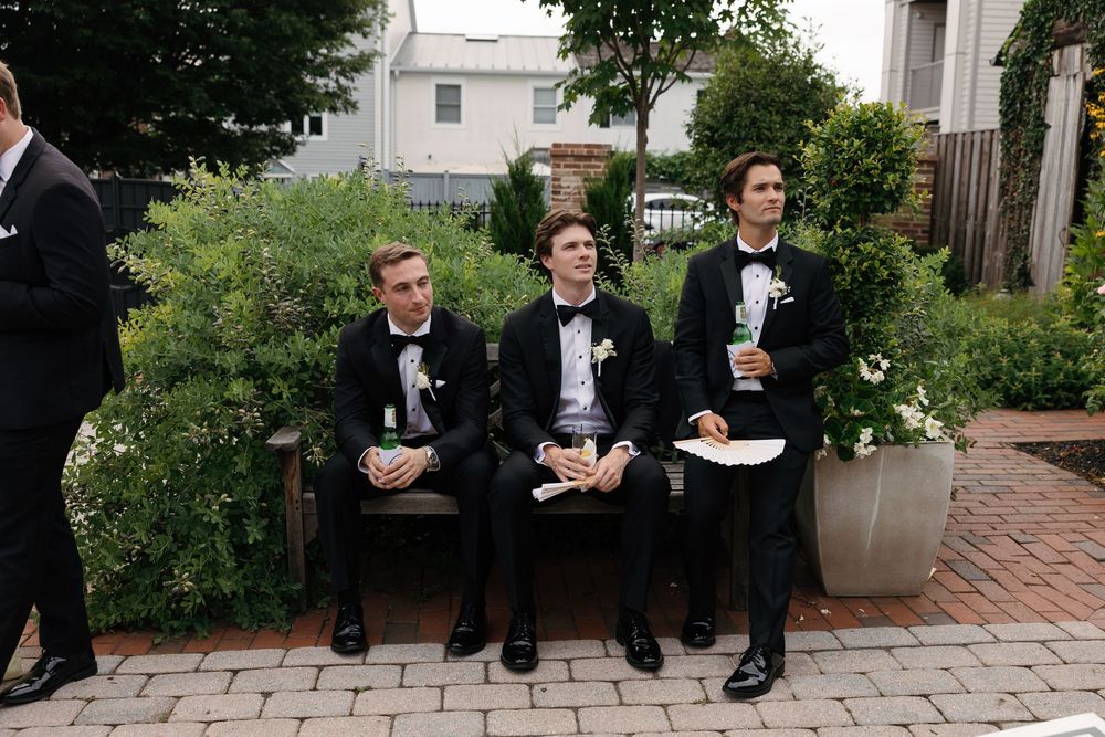 Three groomsmen in black tuxedos with boutonnieres sit on a bench at an outdoor wedding venue.