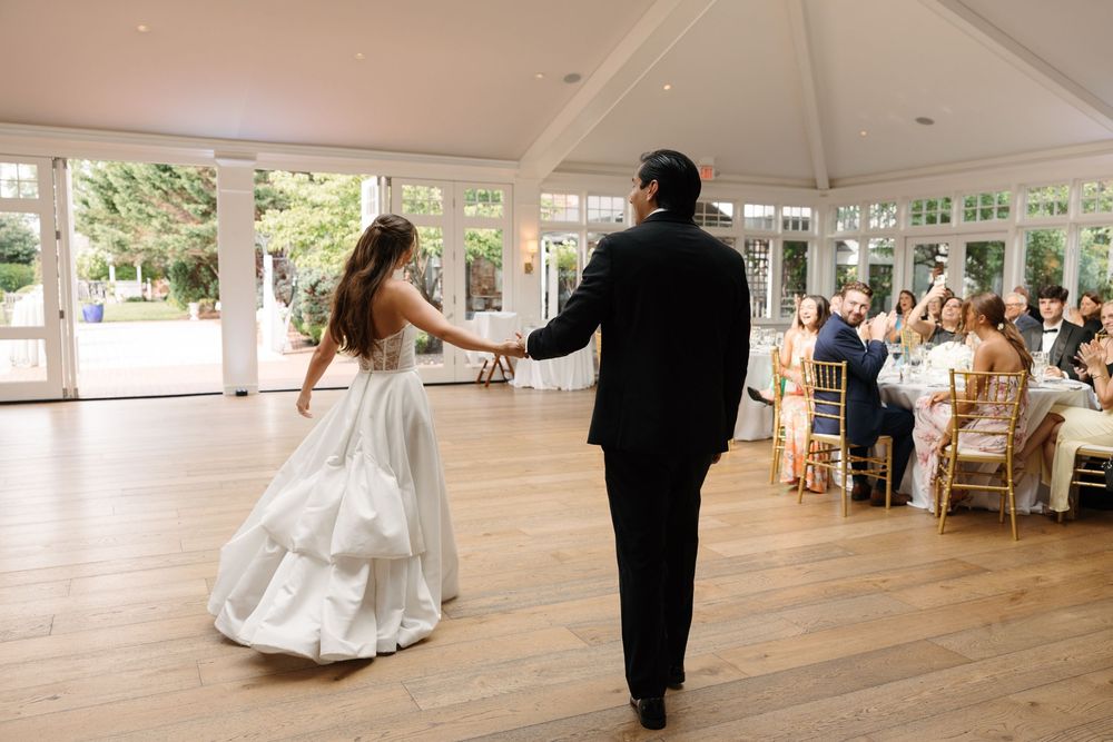 Bride and groom share their first dance on a wooden floor in an elegant glass-windowed wedding reception venue.