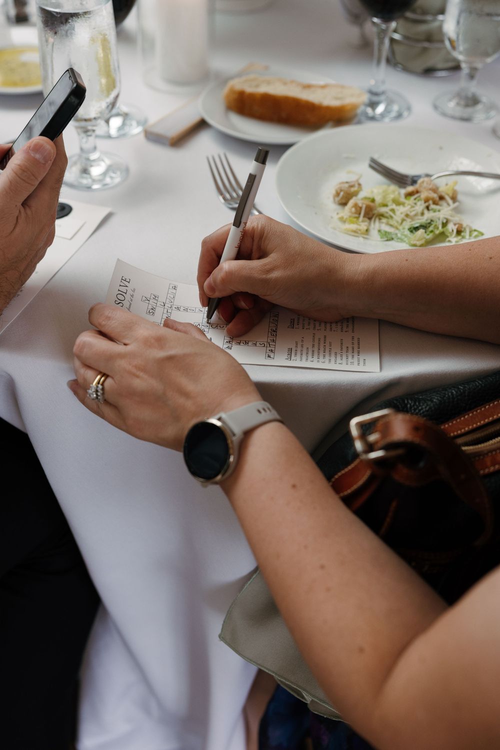 Hands signing a restaurant bill at a dining table with a Caesar salad, bread, and wine glasses nearby.