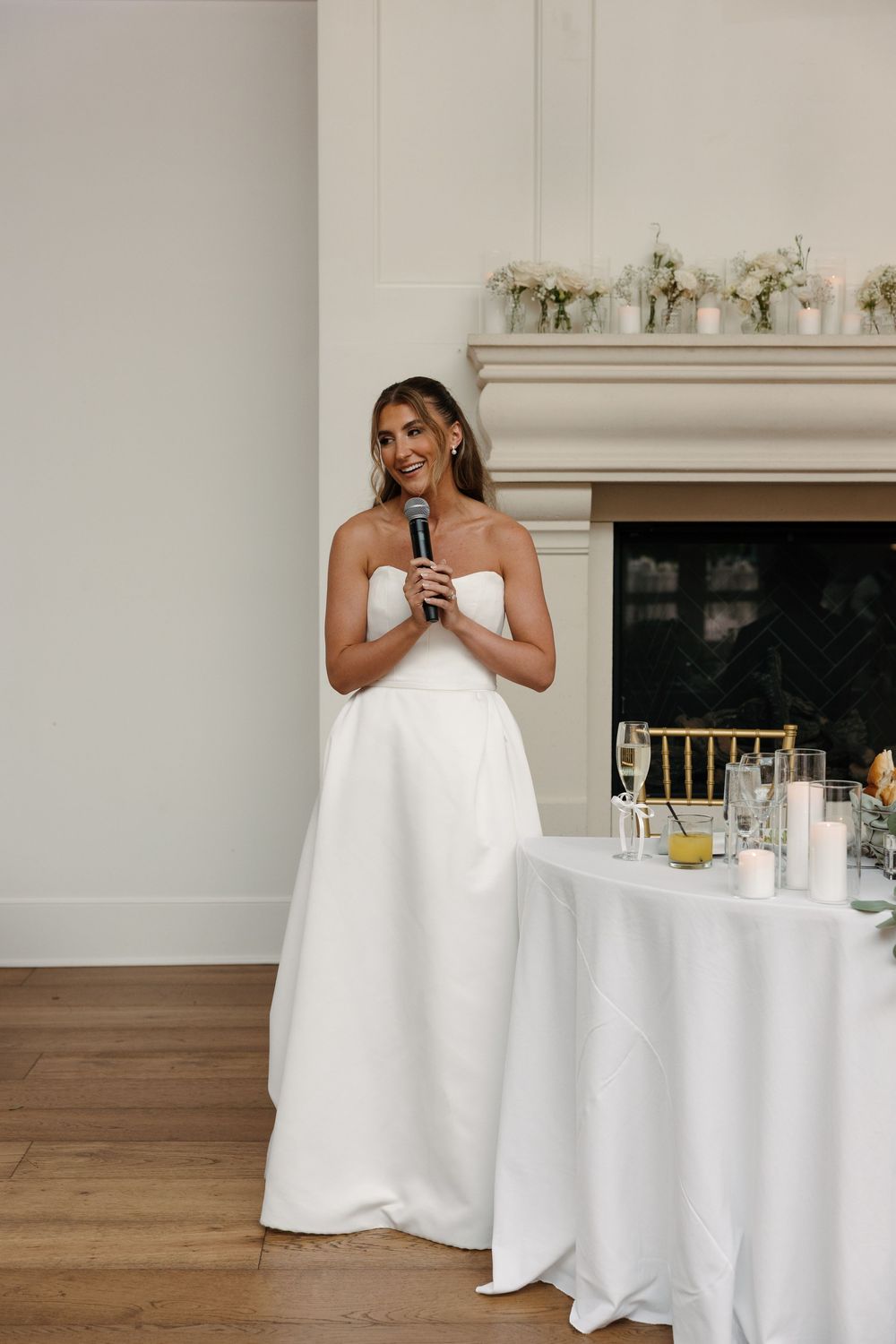 Bride in strapless white gown holding microphone giving speech at elegant wedding reception.