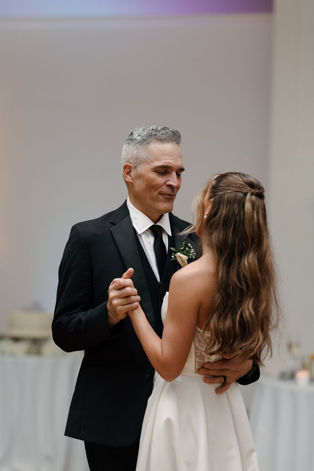 Father and bride share an emotional first dance at wedding reception, man in black suit with boutonniere.