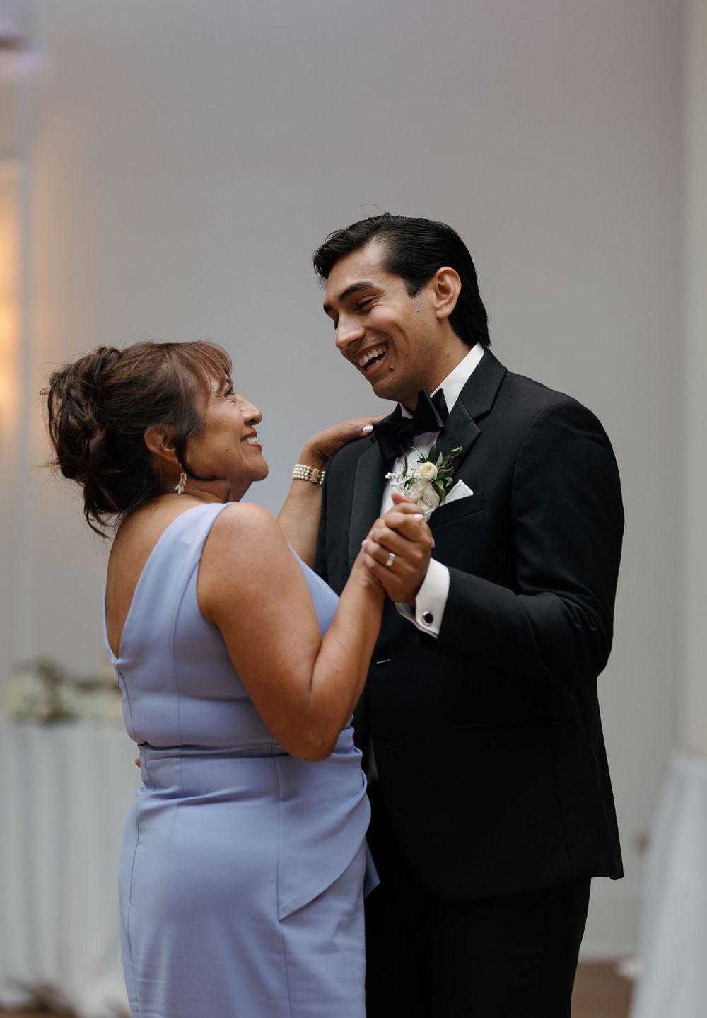 A smiling groom in a black tuxedo shares a mother-son dance at a wedding reception.