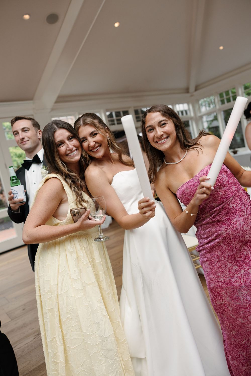 Three women in formal gowns smile at a wedding reception, with a groom in a tuxedo visible in the background.